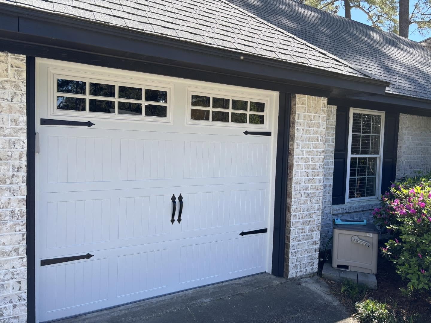 A white garage door with black decorative hardware and small windows at the top, set in a brick house with a window and some landscaping on the side.