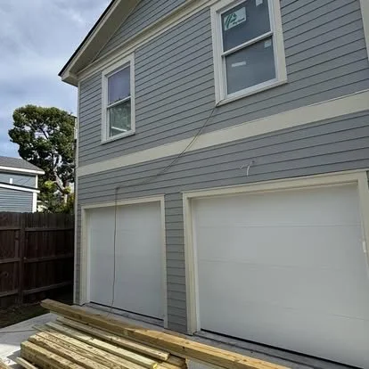 Side of a house with two closed garage doors, grey siding, and two windows on the upper floor. Piles of wood are on the ground in front of the garage doors.