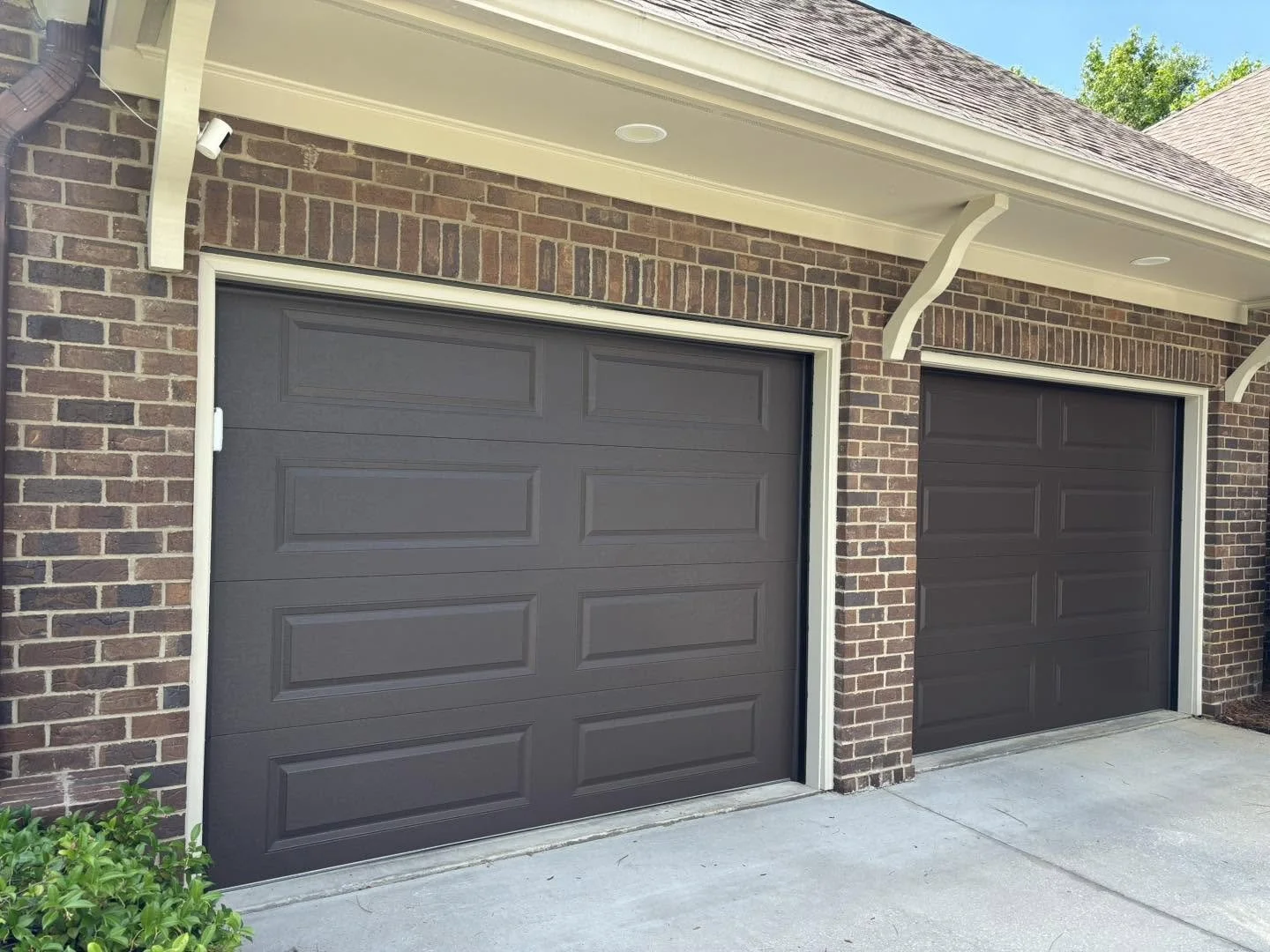 Two closed brown garage doors on a brick house, with white trim, under a white overhang, and a small green bush in the lower left corner.