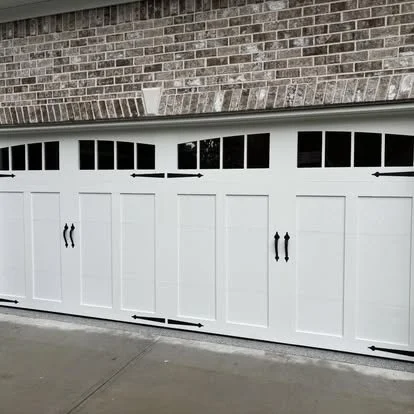 A white garage door with black handles and decorative hardware, set in a brick house wall.