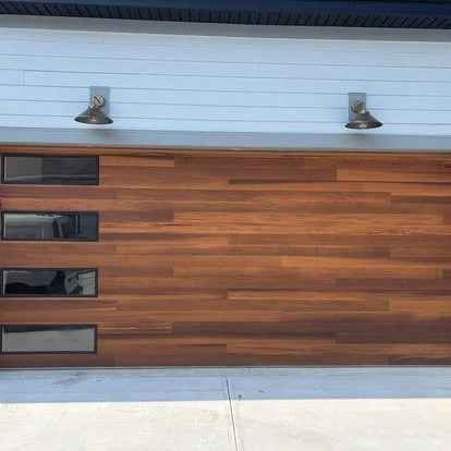 Modern garage door with horizontal wooden panels and small rectangular windows, flanked by two outdoor wall-mounted lights.