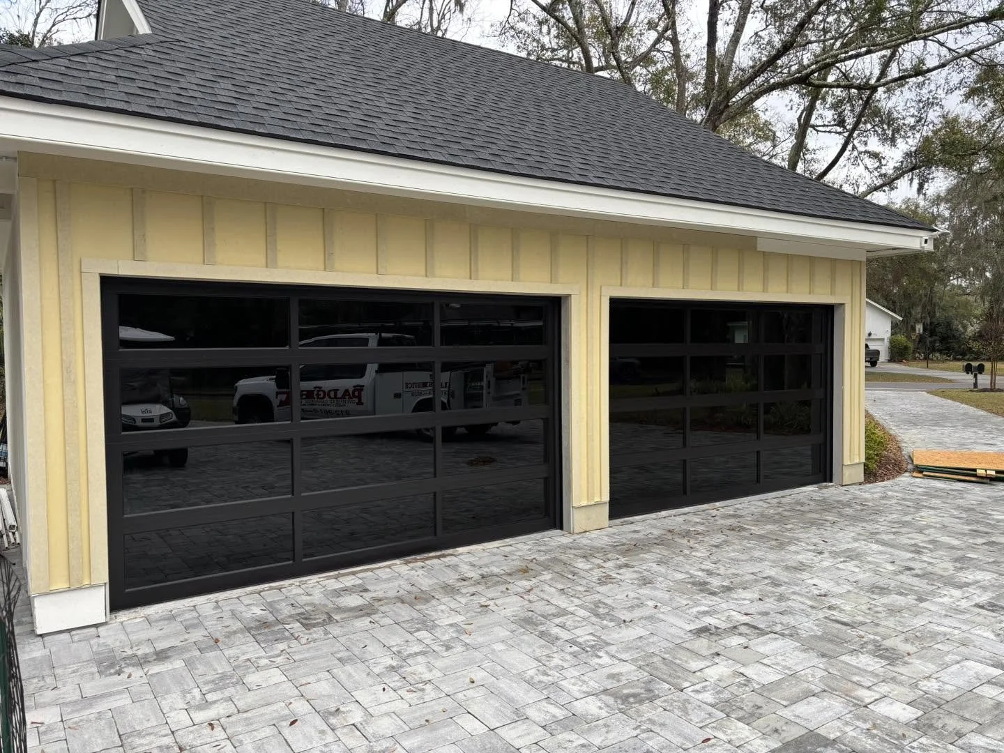 Newly constructed house with two black garage doors, yellow siding, paving stone driveway, and mature trees in the background.