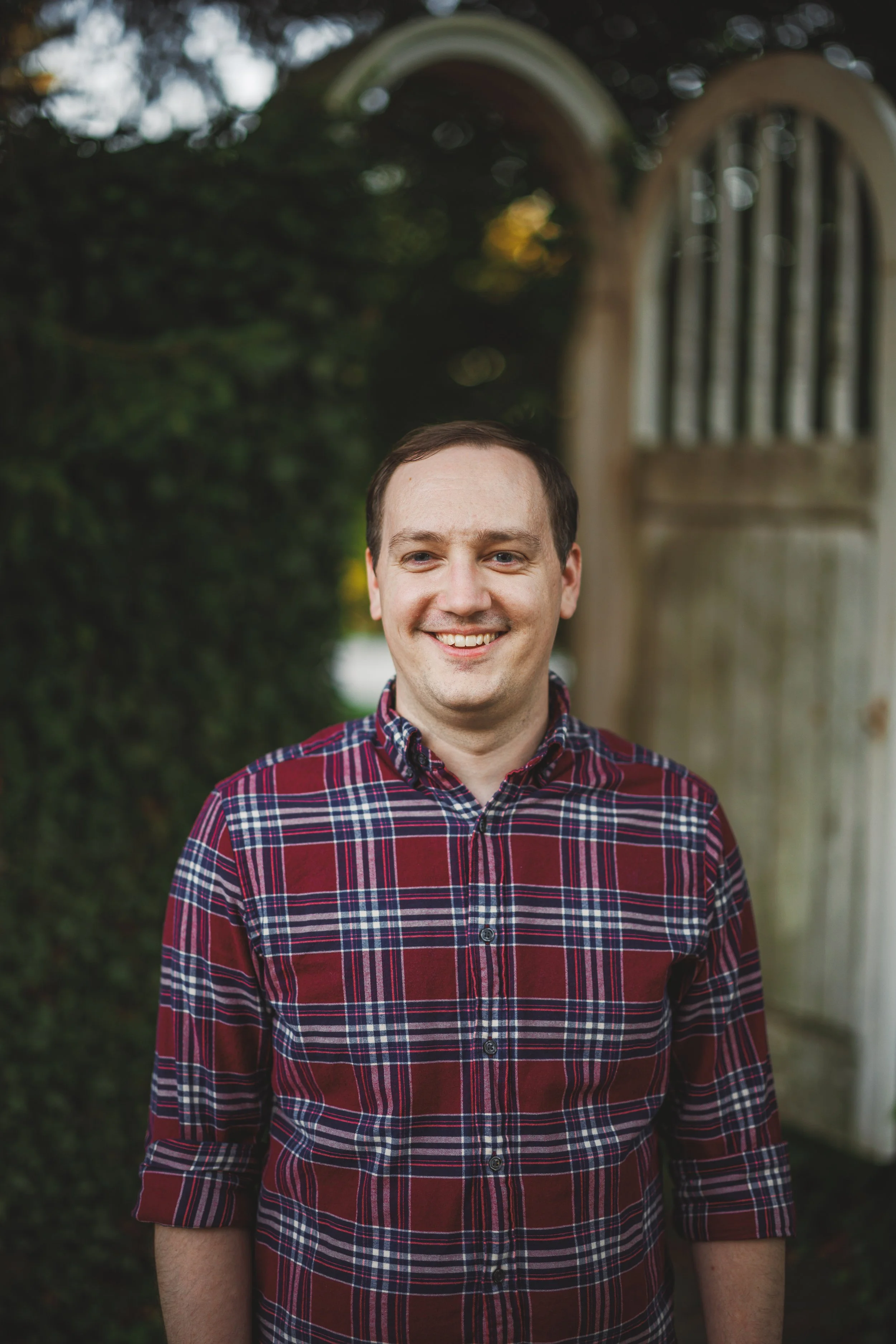 A smiling young man wearing a red, blue, and white plaid shirt standing outdoors during daytime, with greenery and a wooden structure in the background.