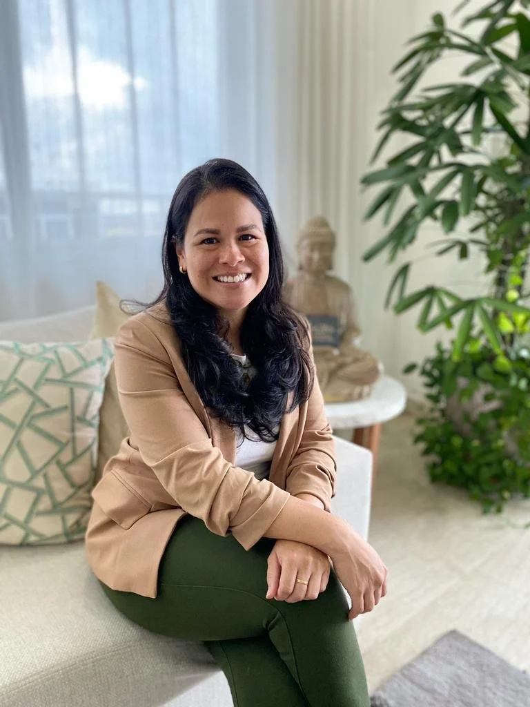 A woman sitting on a beige couch in a bright room with large windows, wearing a tan blazer and dark green pants, smiling and looking at the camera, with plants and a decorative sculpture in the background.