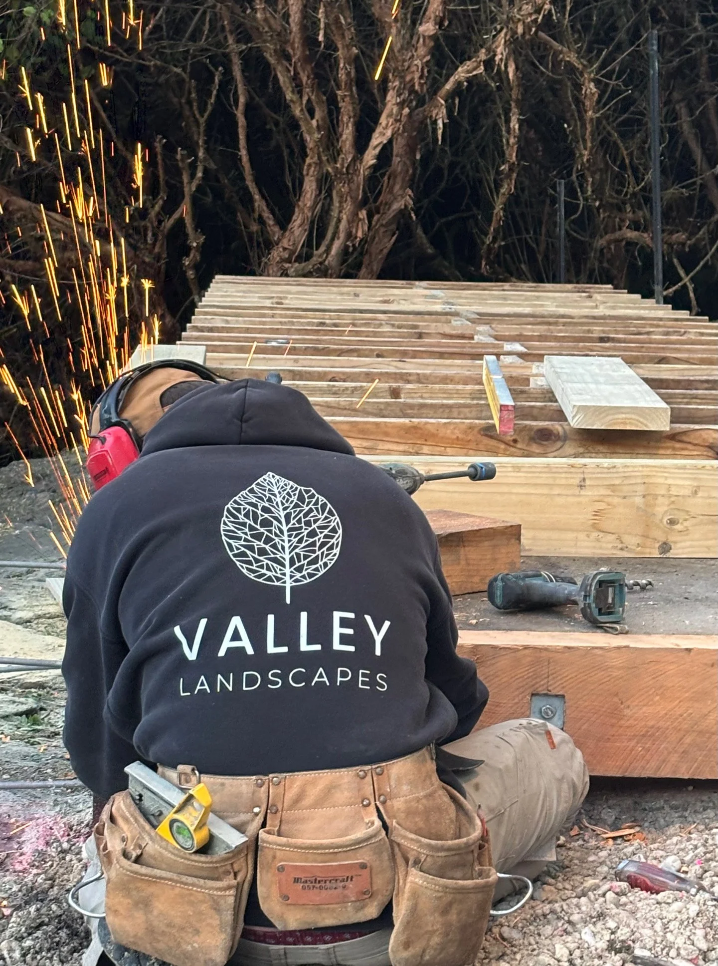 Construction worker kneeling on dirt with a tool belt, working on a wooden deck with a power drill nearby, and sparks flying from a welding process, with a tree in the background.