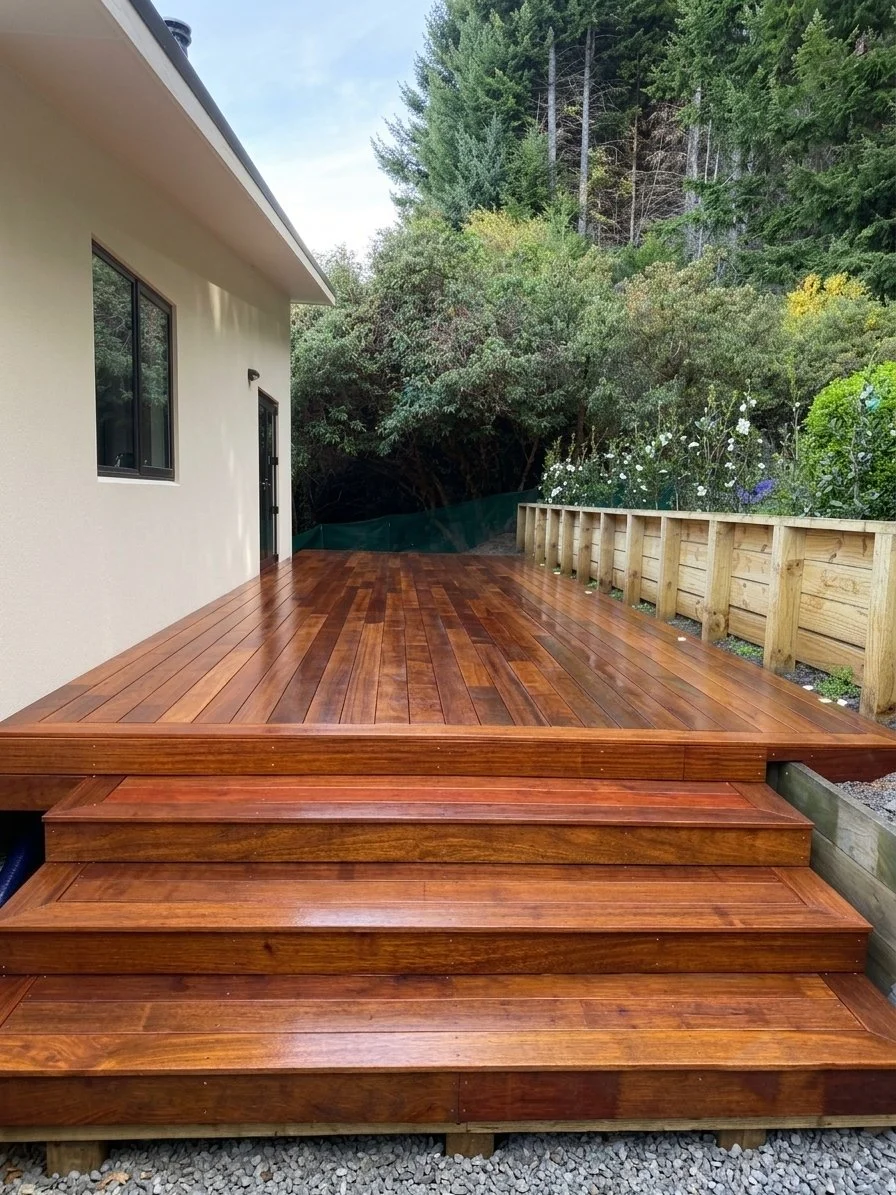 Backyard with a newly built wooden deck, steps leading up to the deck, adjacent house with window and door, surrounding lush trees and plants.