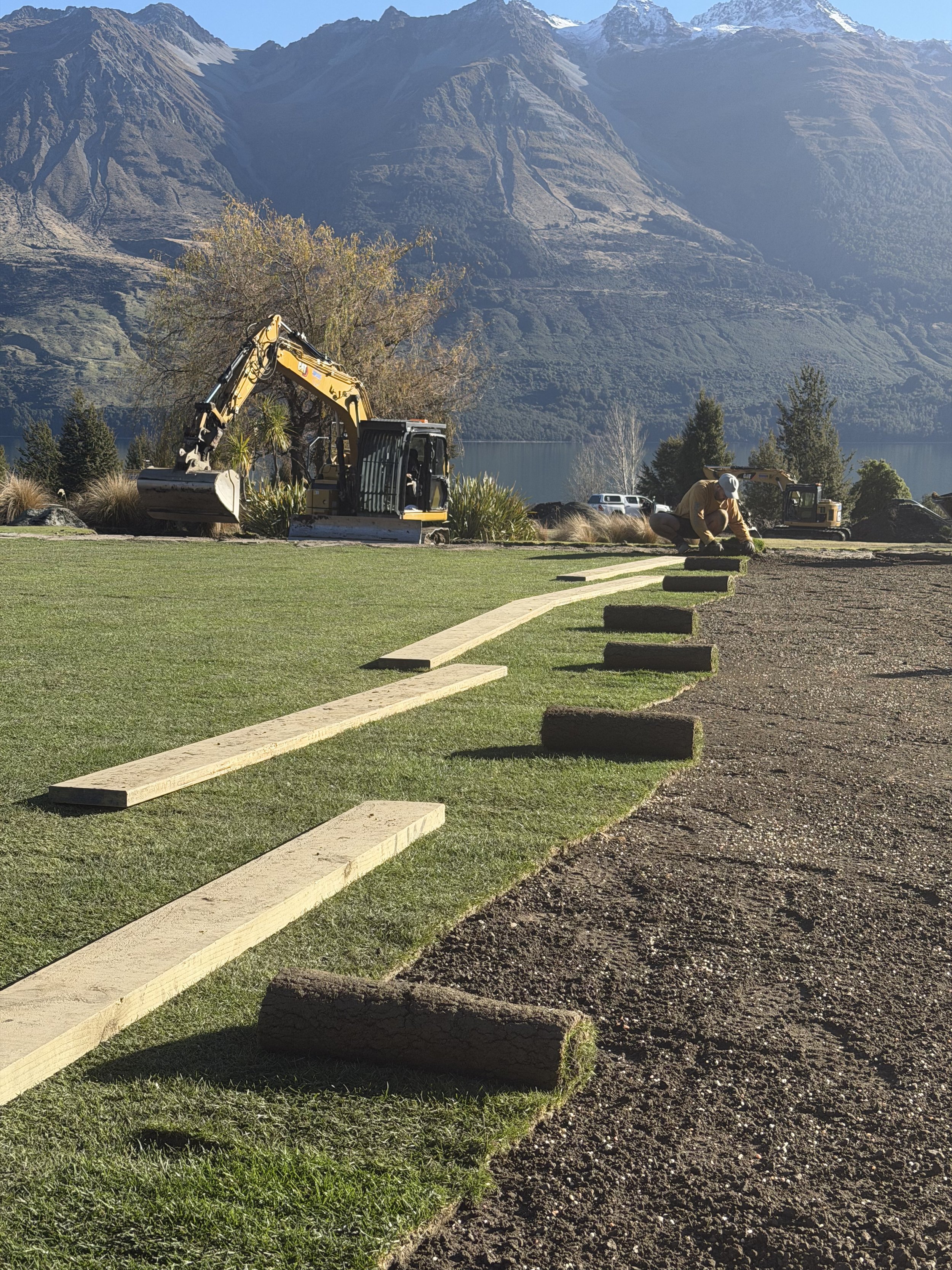 A person installing a border of wooden logs along a landscaped yard with grass and trees, with construction equipment and mountains in the background.