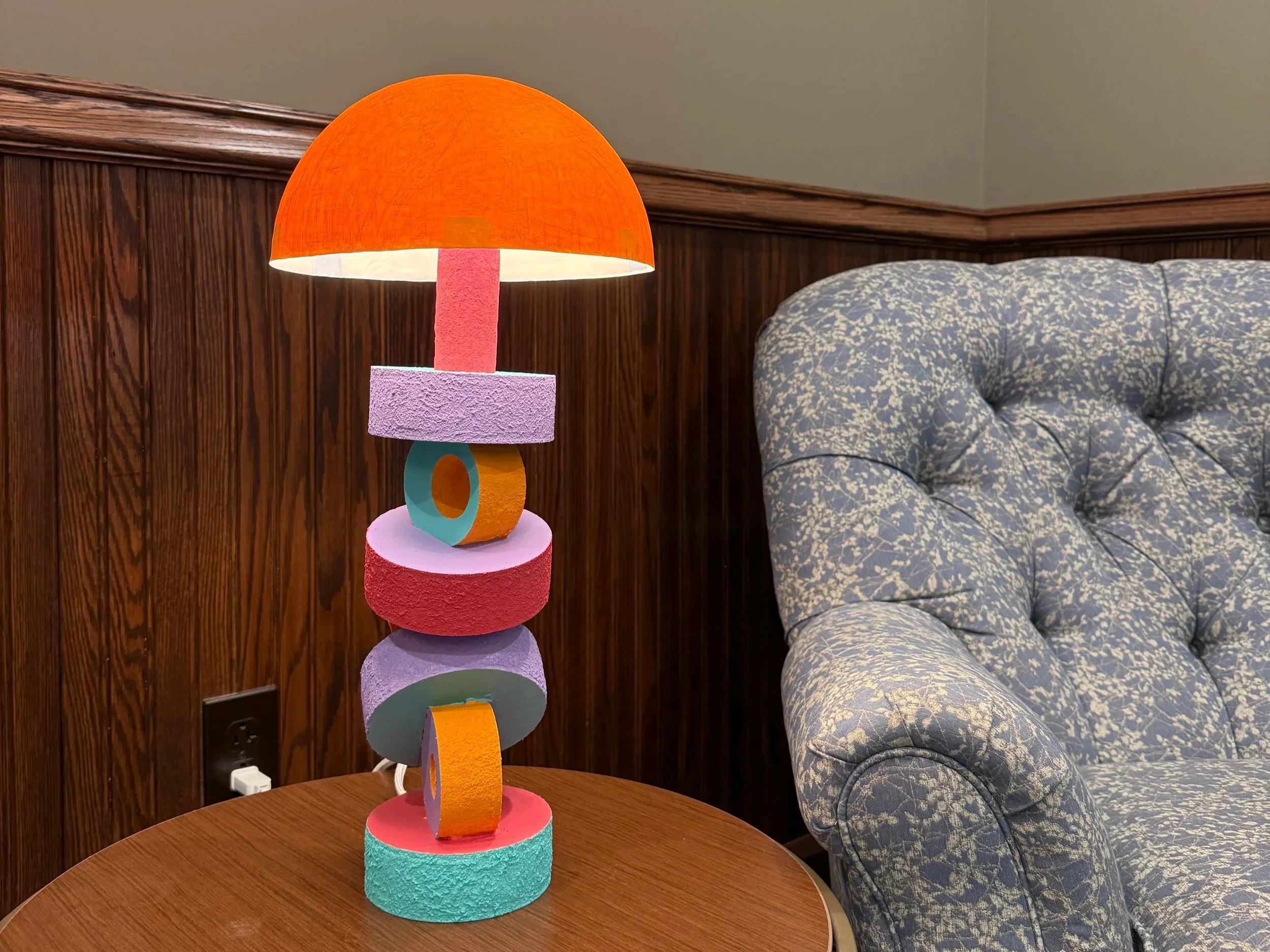 Colorful abstract sculpture on a wooden table, next to a blue patterned sofa and dark wood wall paneling.