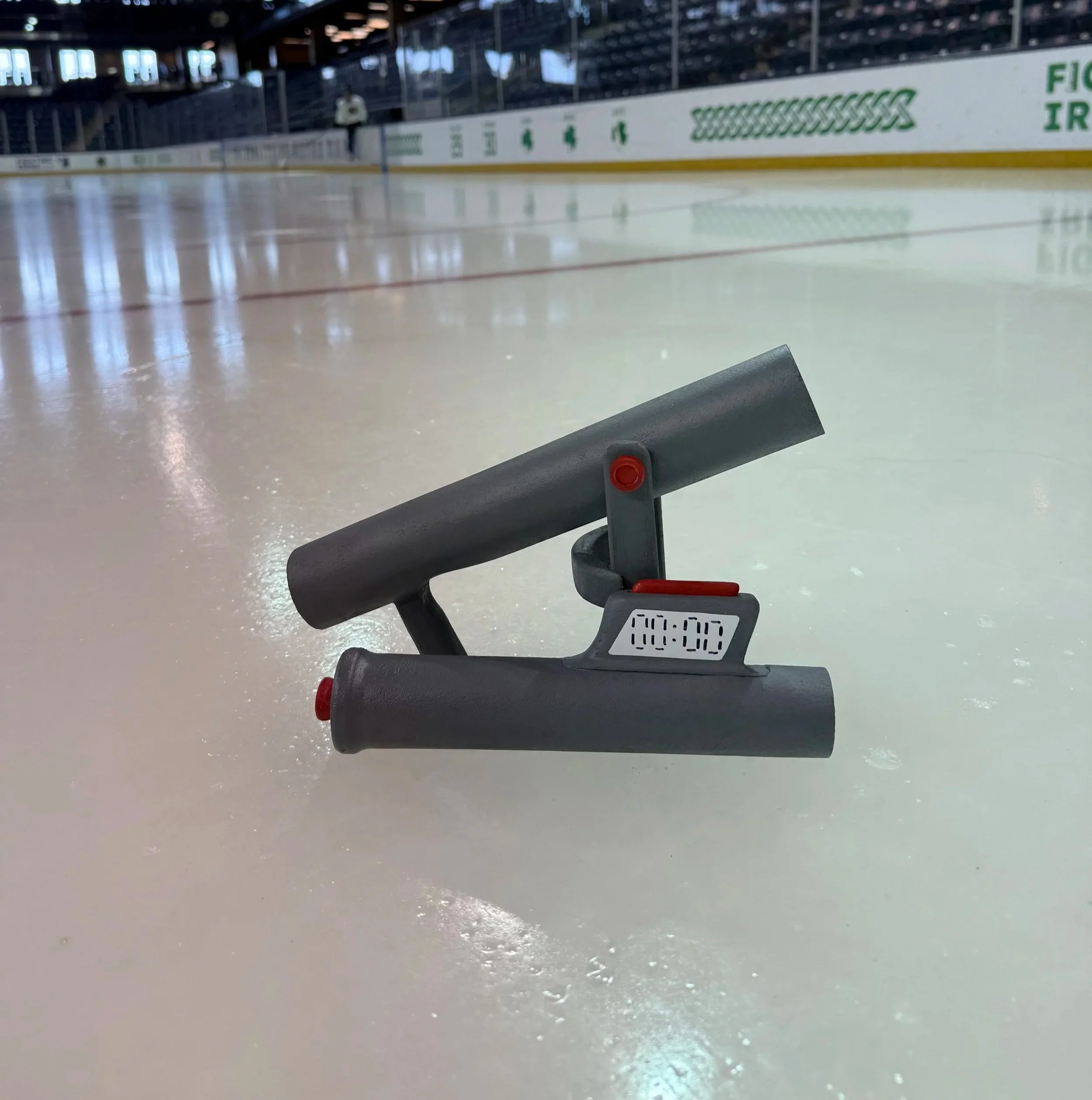 An ice rink with a skating scoreboard and a person on the distant side of the rink, blurred in the background. The photo is taken from the perspective of someone looking down at a skating aid or barrier, with a scoreboard displaying time in the foreground.