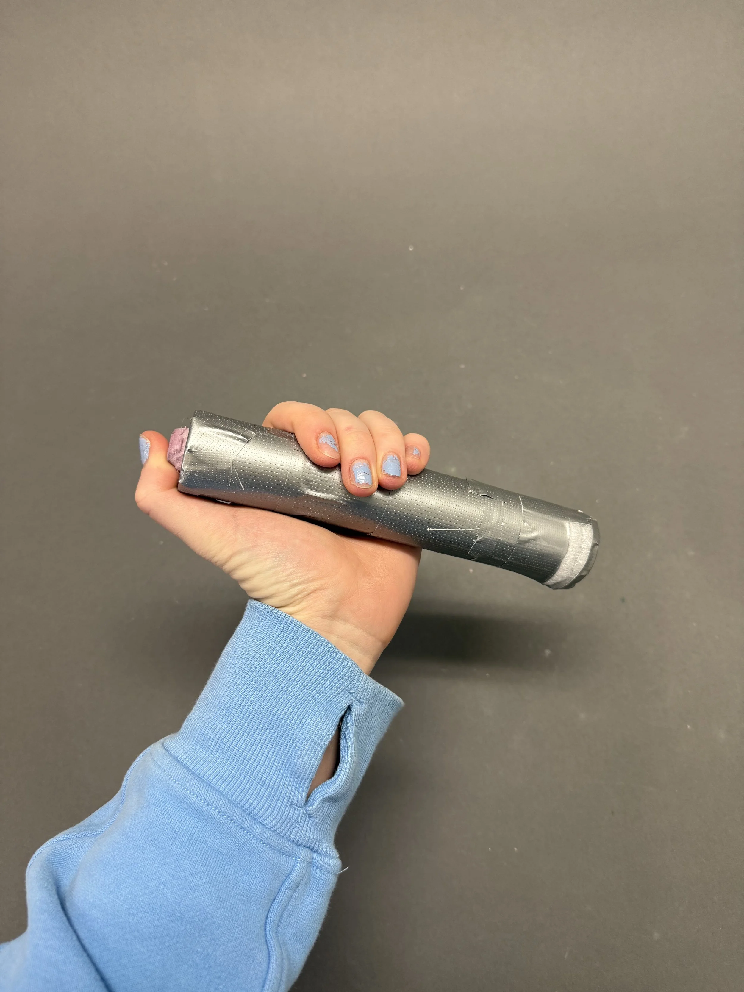 A person with painted nails holding a cylindrical object wrapped in gray tape, wearing a light blue long sleeve shirt, against a dark gray background.