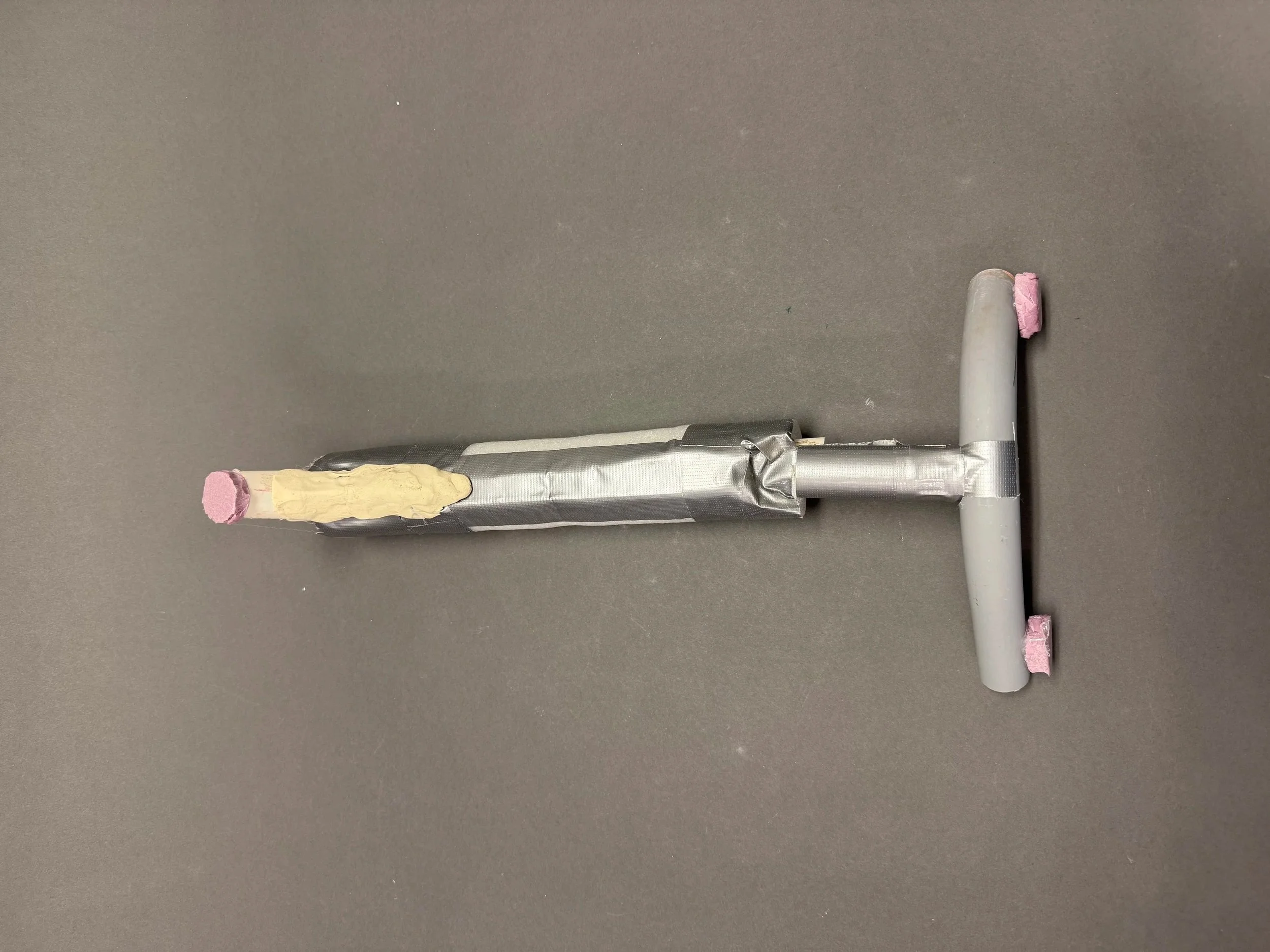 A gray cylindrical object wrapped in silver foil with pink foam padding at both ends, connected to two white cardboard tubes with pink foam padding, set against a plain gray background.