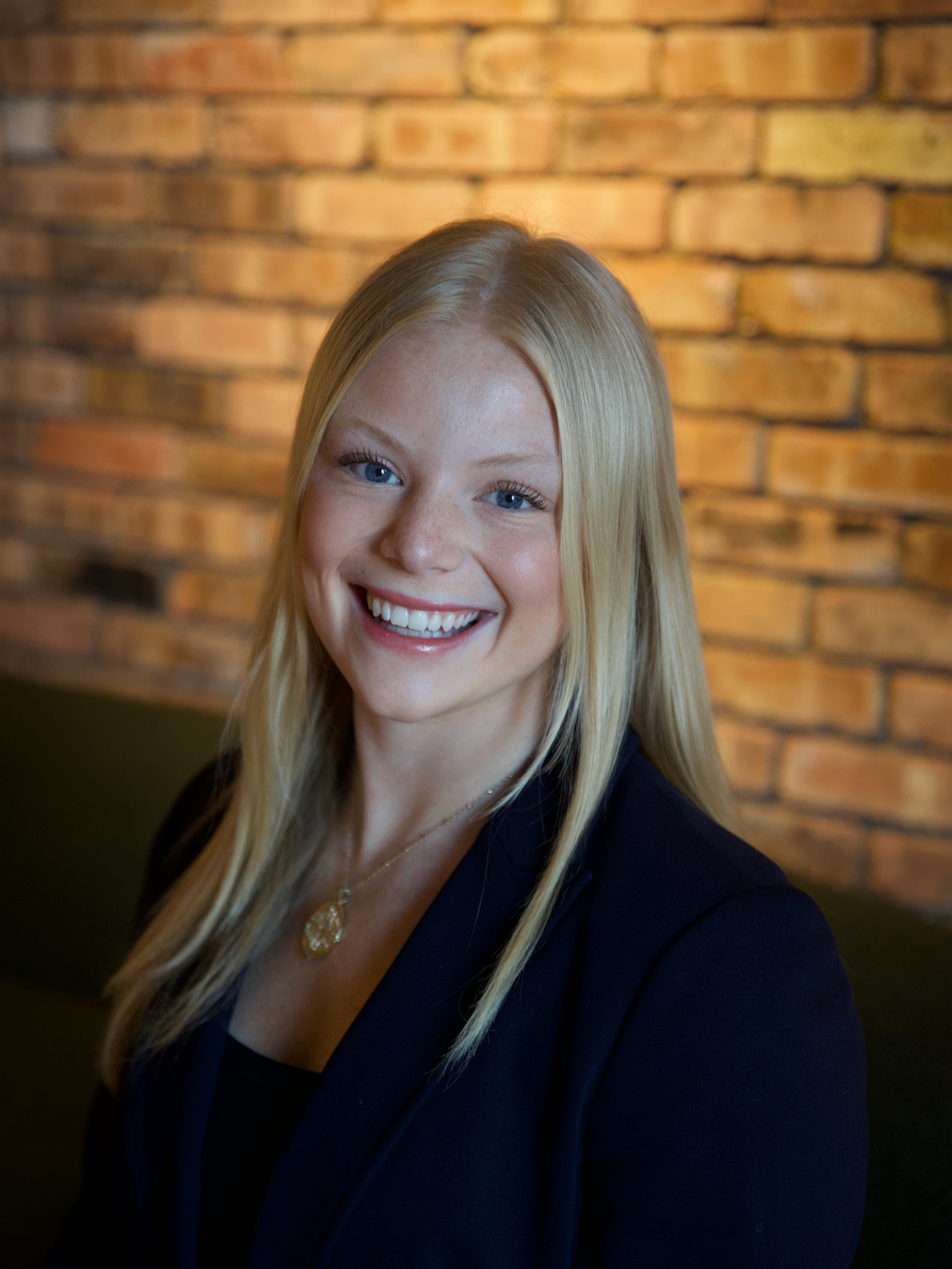 A smiling young woman with long blonde hair, wearing a black blazer and gold necklace, standing in front of a brick wall.