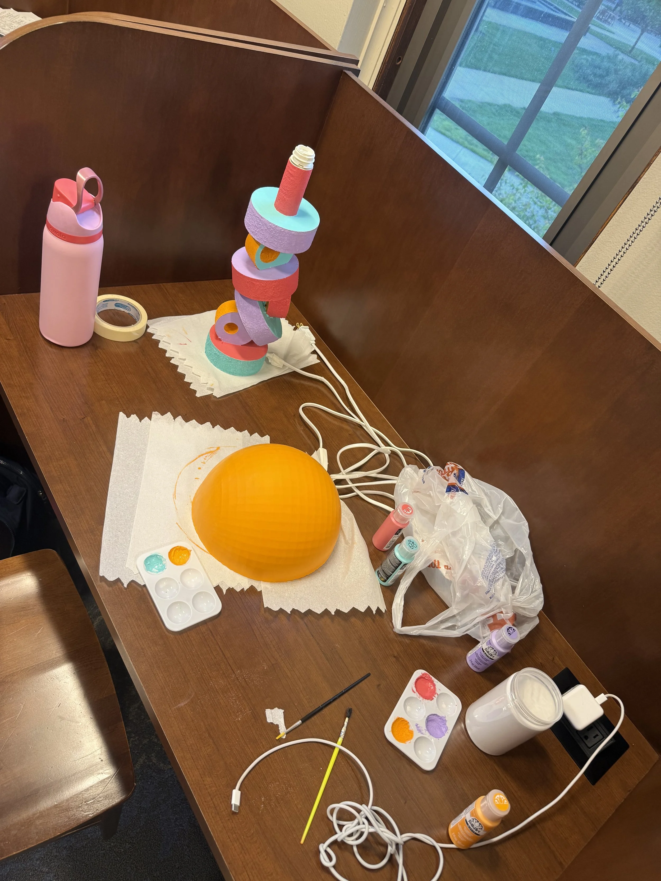 A wooden table with art supplies, including a stack of colorful foam rings, a yellow paintbrush, paint palettes with red, purple, orange, and teal paints, a pink water bottle, a large orange paper lantern, various paint bottles, a paint tube, brushes, and electrical cords. There is lace paper towel underneath the foam rings and some scattered paper towels on the table. A window showing greenery and a screened door is in the background.