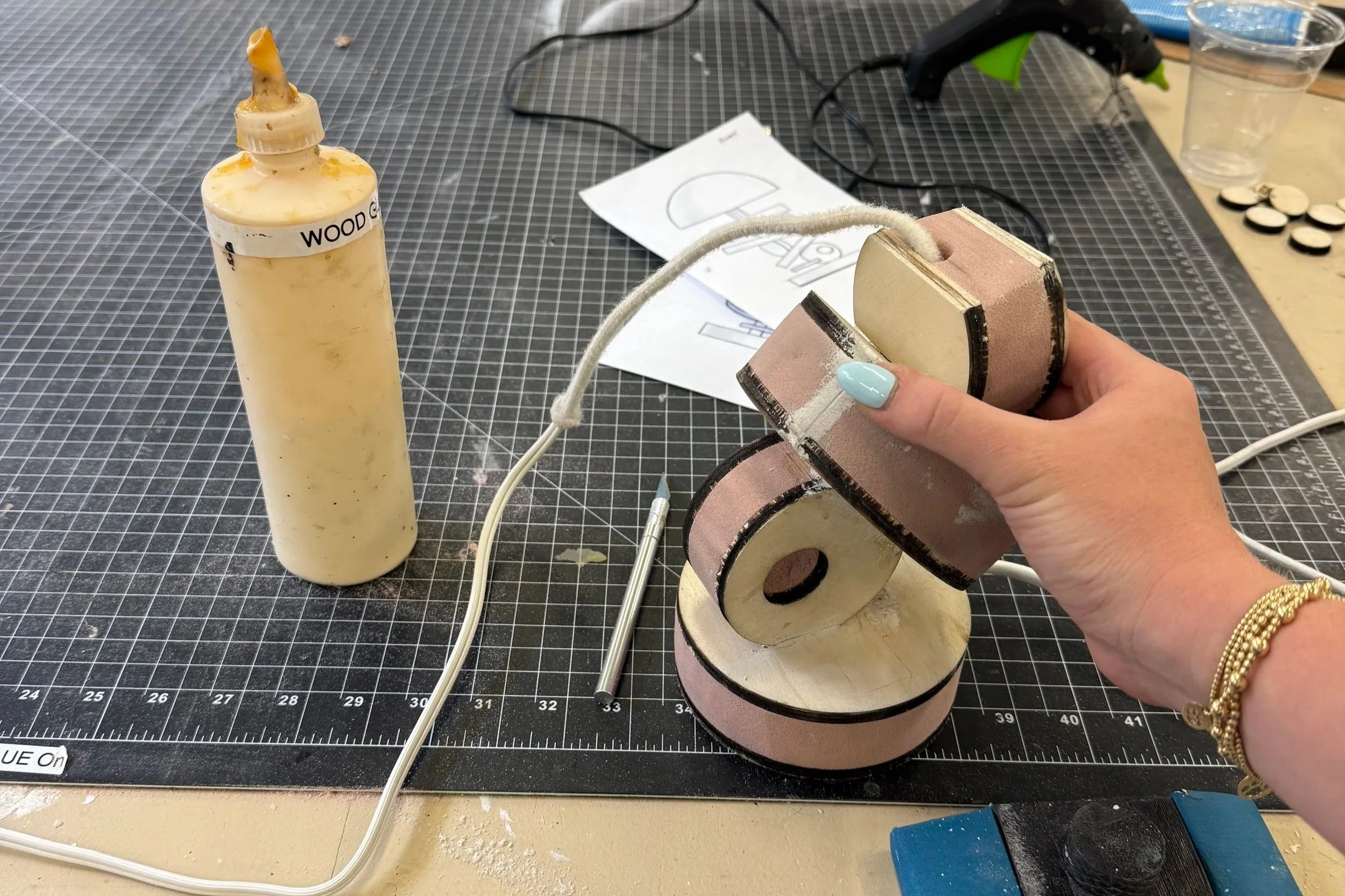 A workspace with craft supplies, including a glue bottle labeled 'Wood Glue,' a hot glue gun, a hand holding small foam blocks, and various tools on a cutting mat.