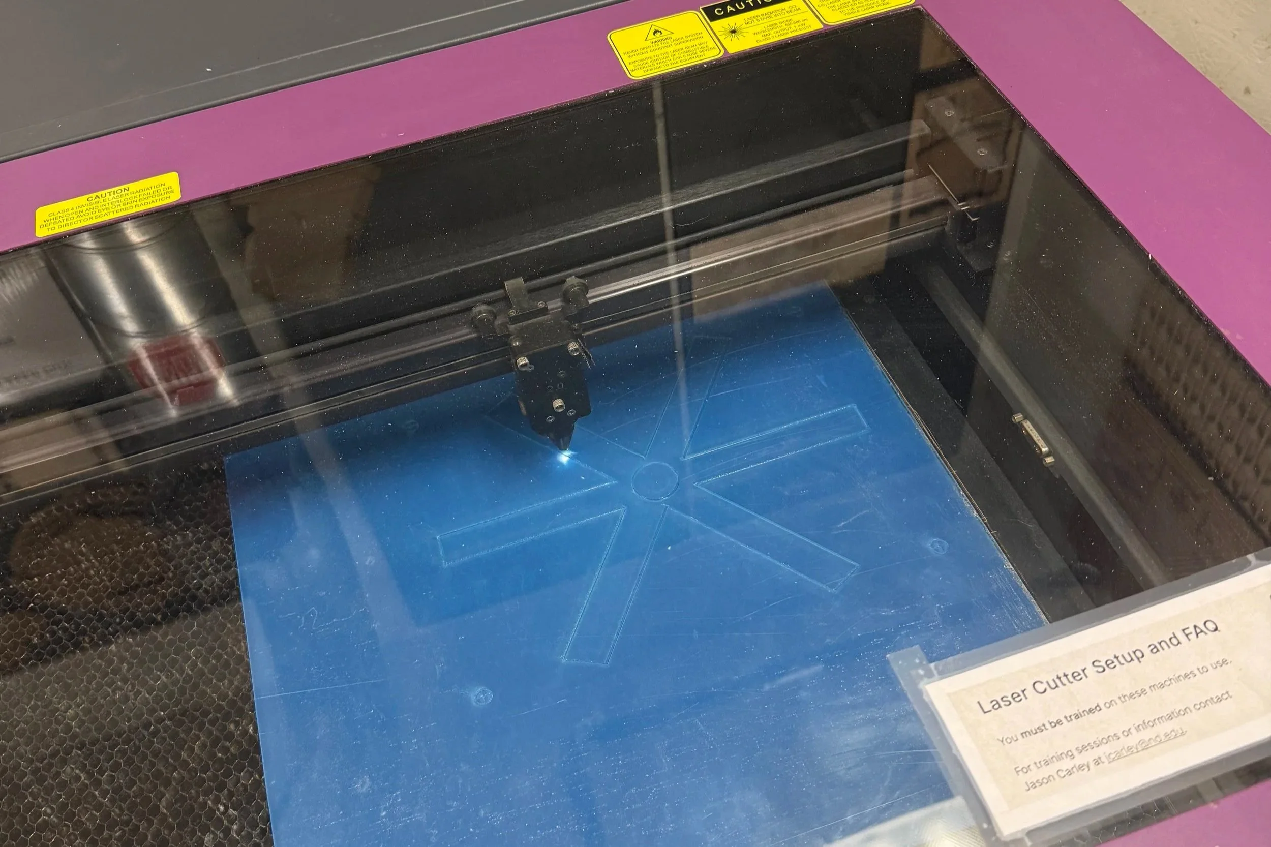A laser cutting machine with a purple top and yellow caution labels, partially visible through a clear window, cutting a blue material with a star pattern.