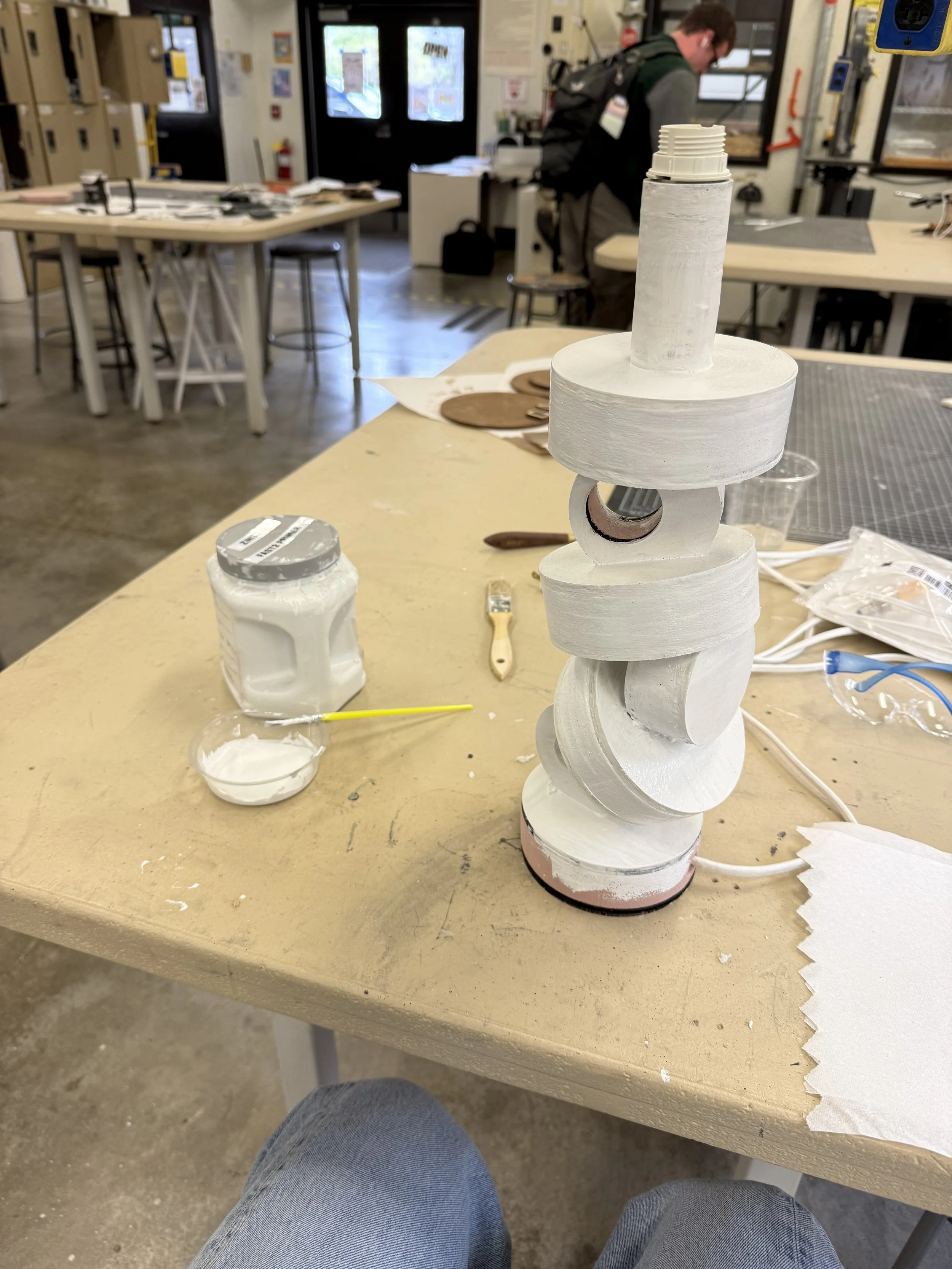 A workshop table with a structure made of white cylindrical objects, some of which are painted white, and tools and supplies around it. In the background, a person is working at a station in a workshop or classroom.
