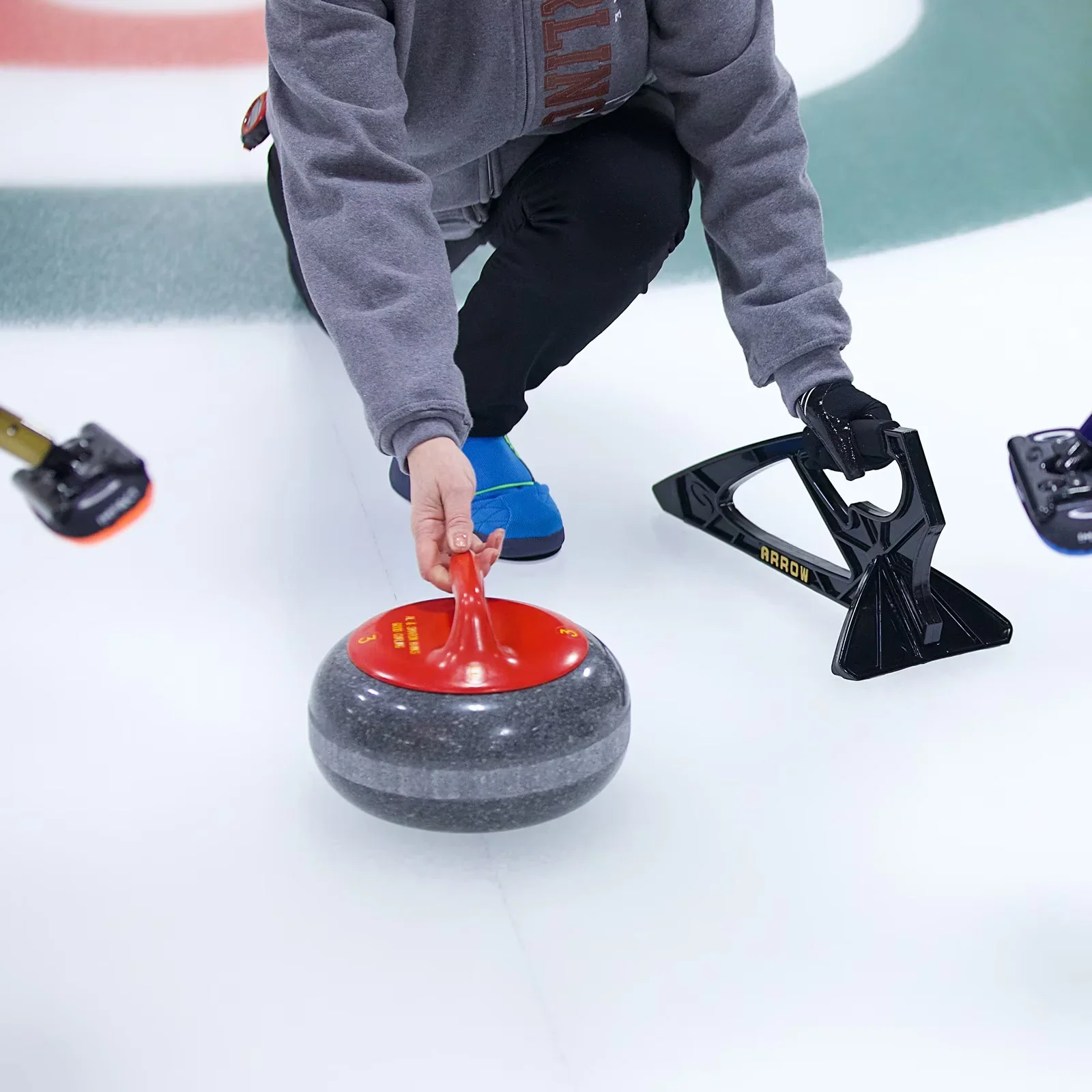A person in a gray hoodie and black pants on an ice rink, preparing to launch a curling stone with a red handle, while holding a curling broom.