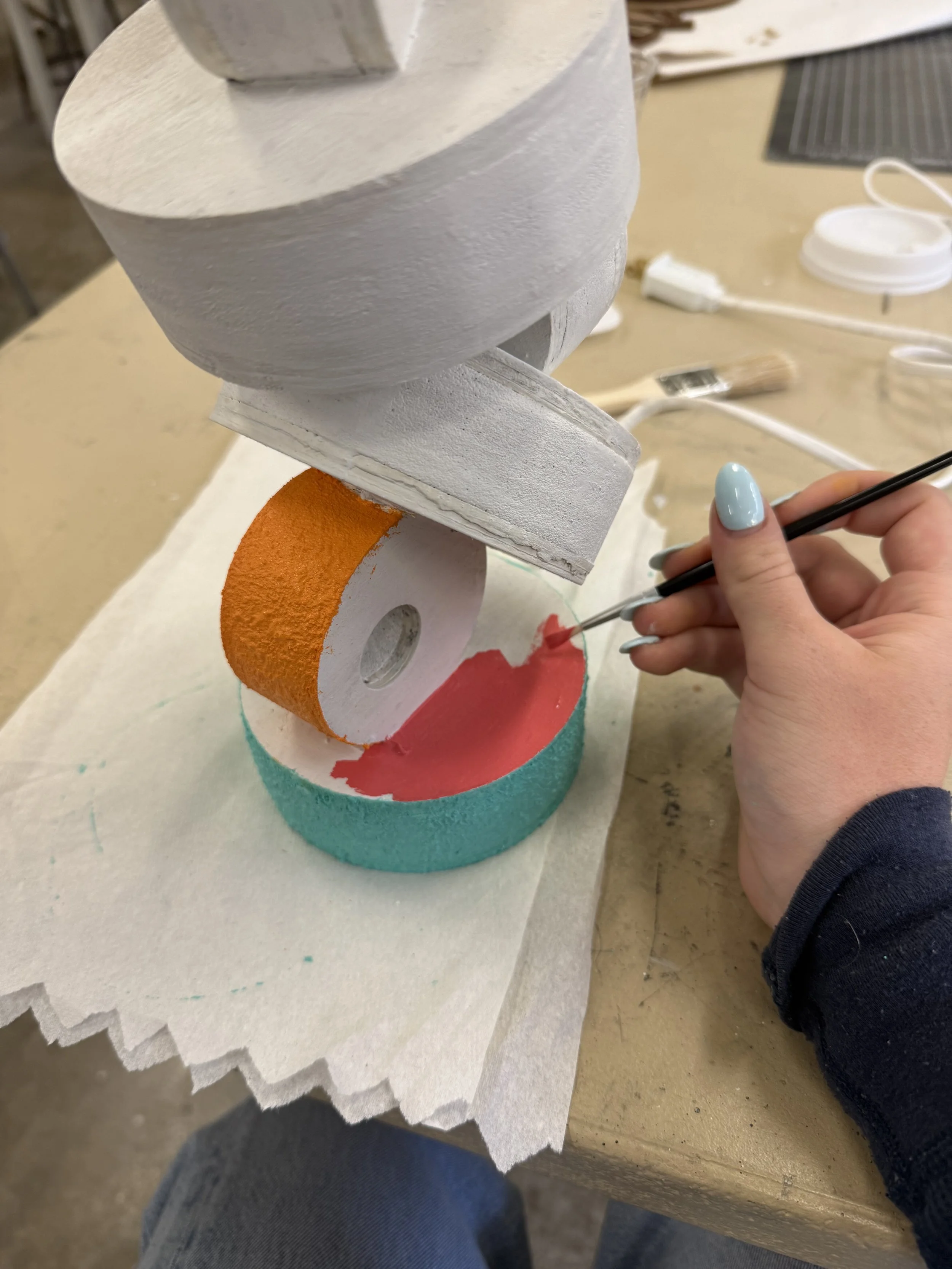 A person painting a large, colorful, abstract sculpture with a small brush. The sculpture features vibrant orange, red, and teal colors and is placed on a table.