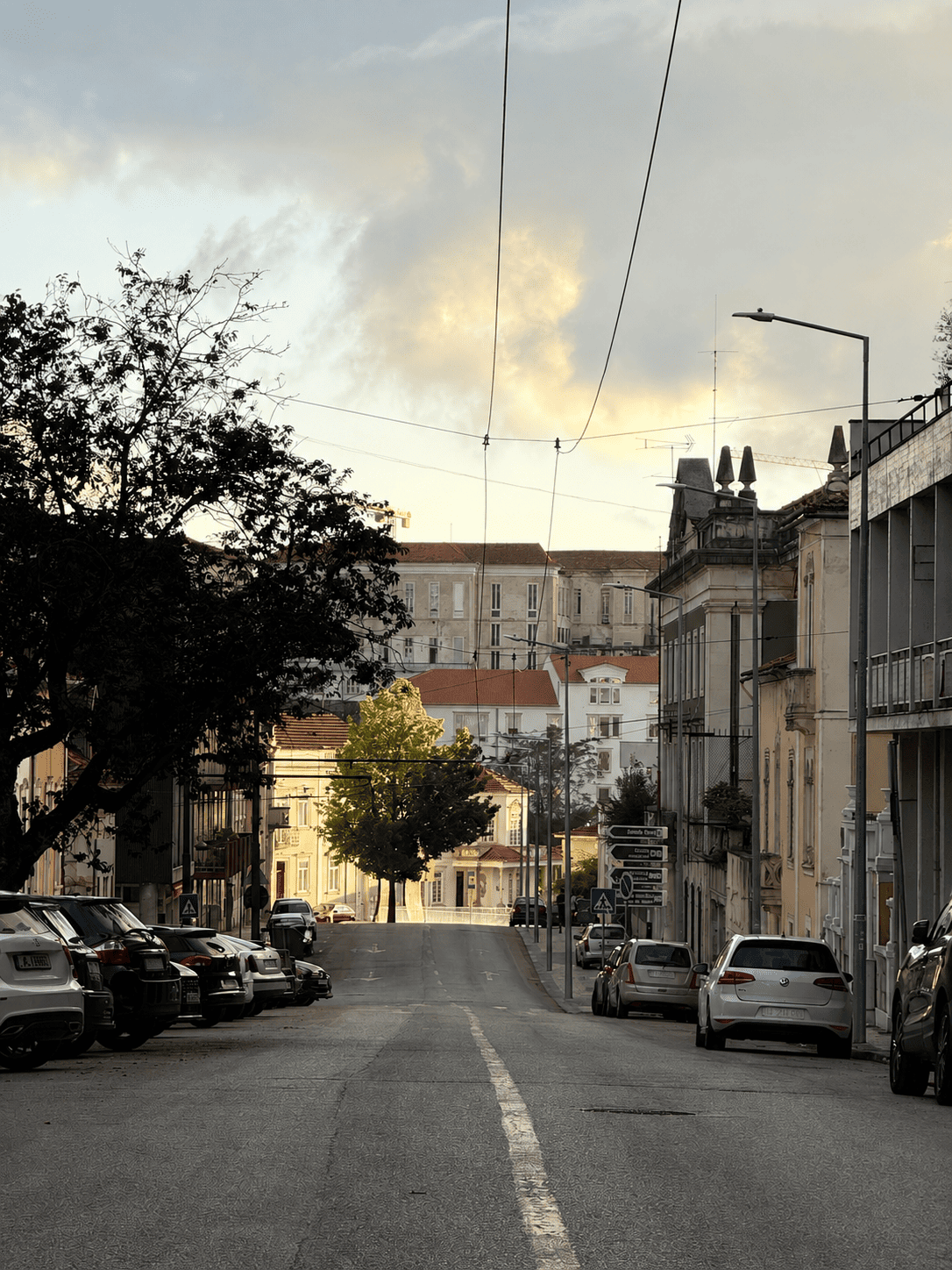 A quiet urban street at dusk with parked cars on both sides, trees lining the sidewalk, multi-story buildings with architectural details, and a sky with clouds illuminated by the setting sun.