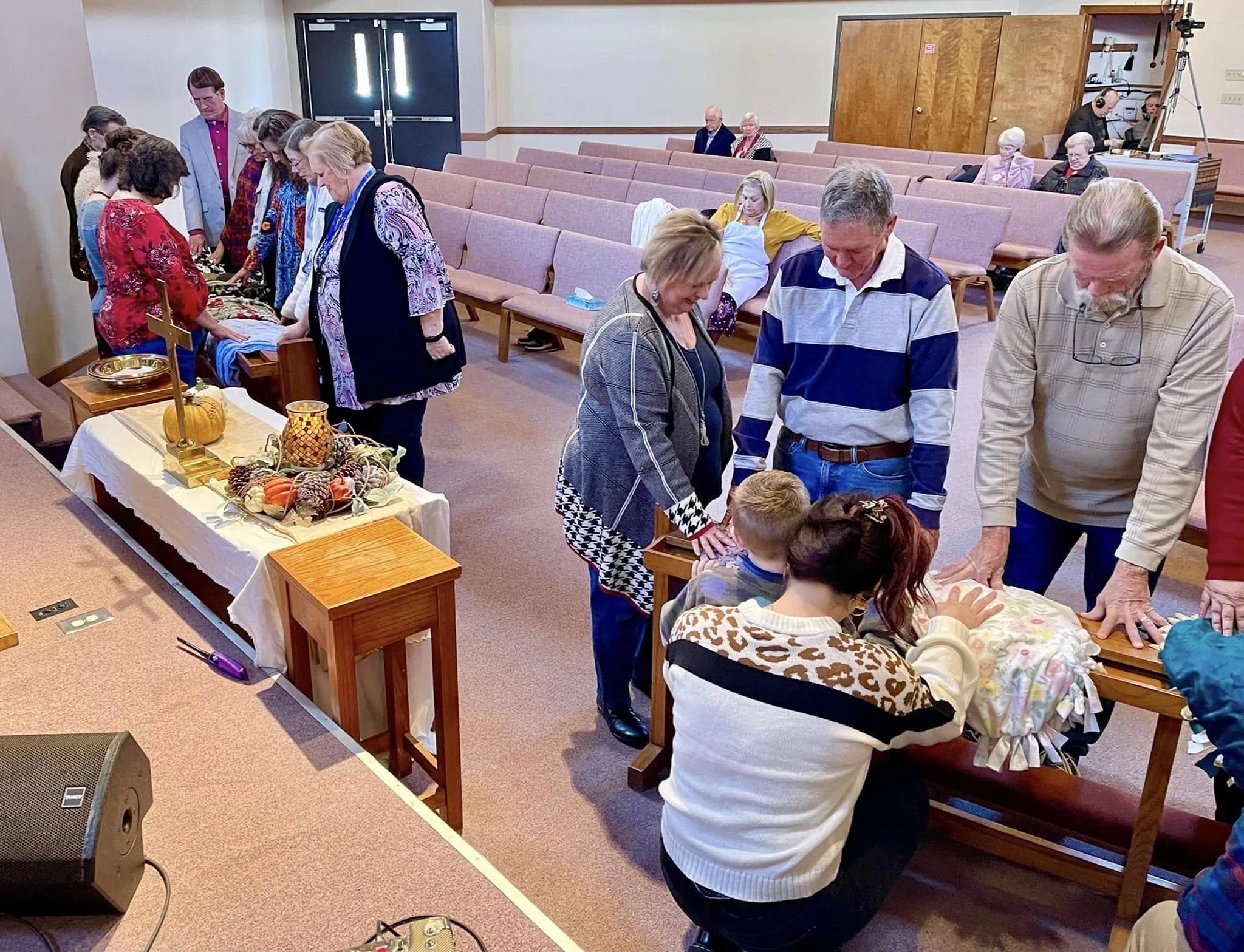 People gathered around tables with colorful cloths, pumpkins, and fall decorations in a church or community center, with some seated in pews or chairs and others standing or leaning over the tables.