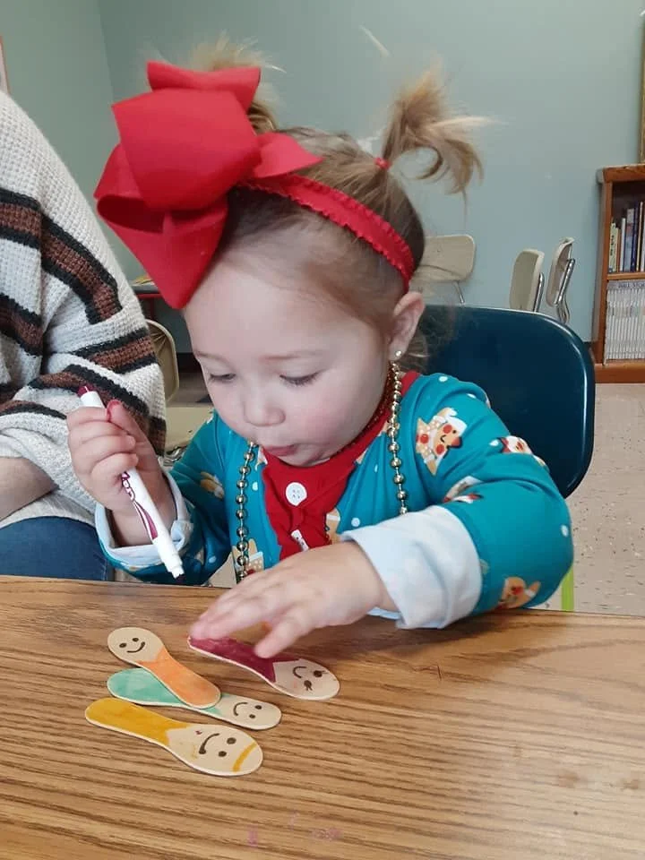A young girl with a large red bow in her hair, wearing a blue pajama with gingerbread men, is sitting at a wooden table. She is painting cutouts of worms with smiley faces and different colors, and holding a marker in her right hand.