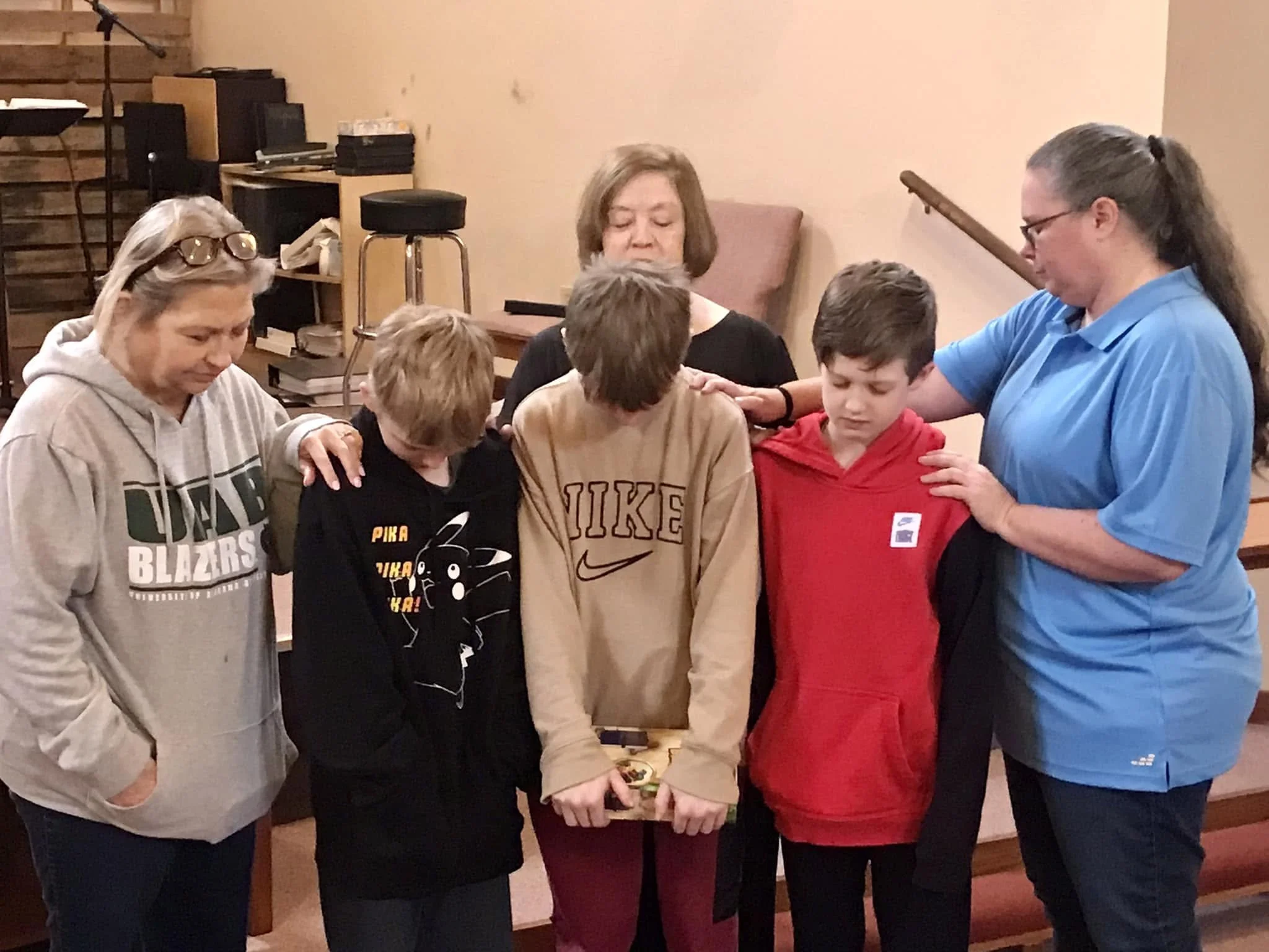 A group of five people, three women and two boys, are gathered around and looking at a small object or device on a table. The setting appears to be an indoor room with a wooden wall and some shelves or cabinets in the background.
