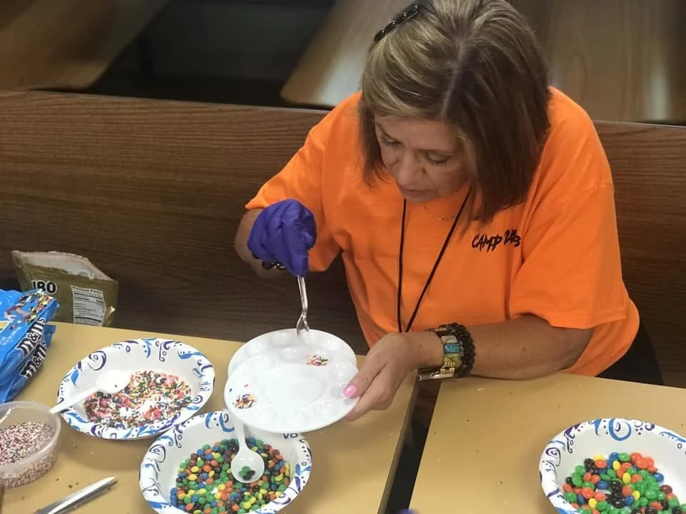 Woman in an orange shirt with 'CAMP' logo, wearing purple glove, decorating a white cake with a fork, surrounded by bowls of colorful sprinkles and candies on a table.