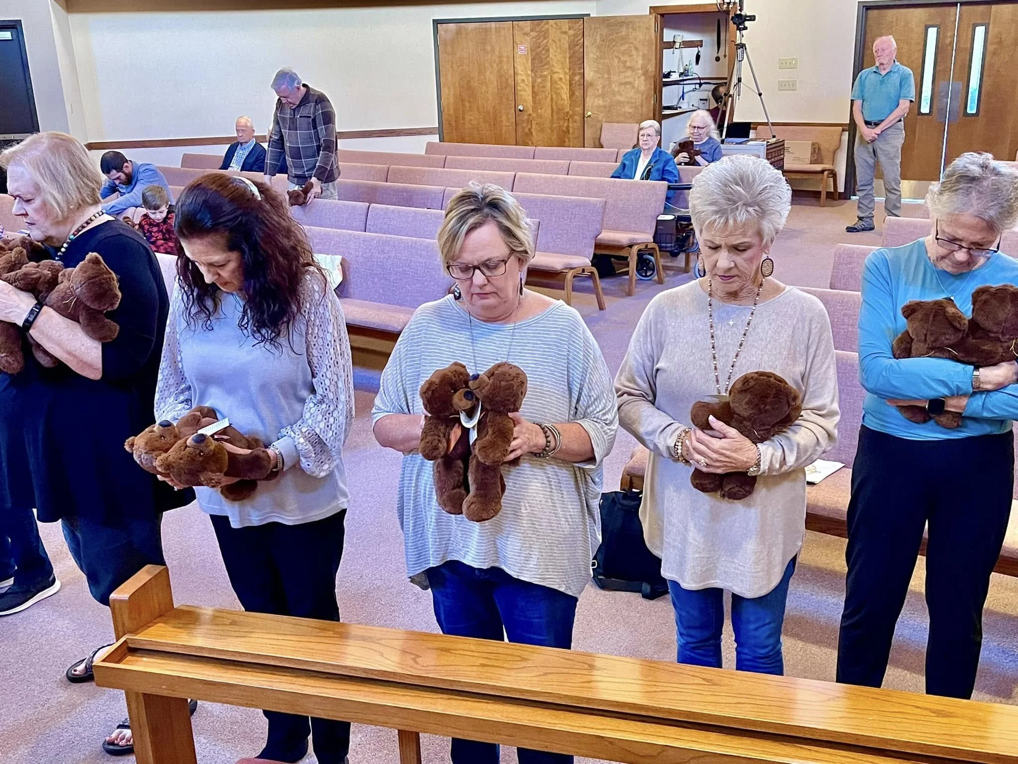 Group of elderly women holding teddy bears, standing in a church or community hall with empty pink chairs behind them.