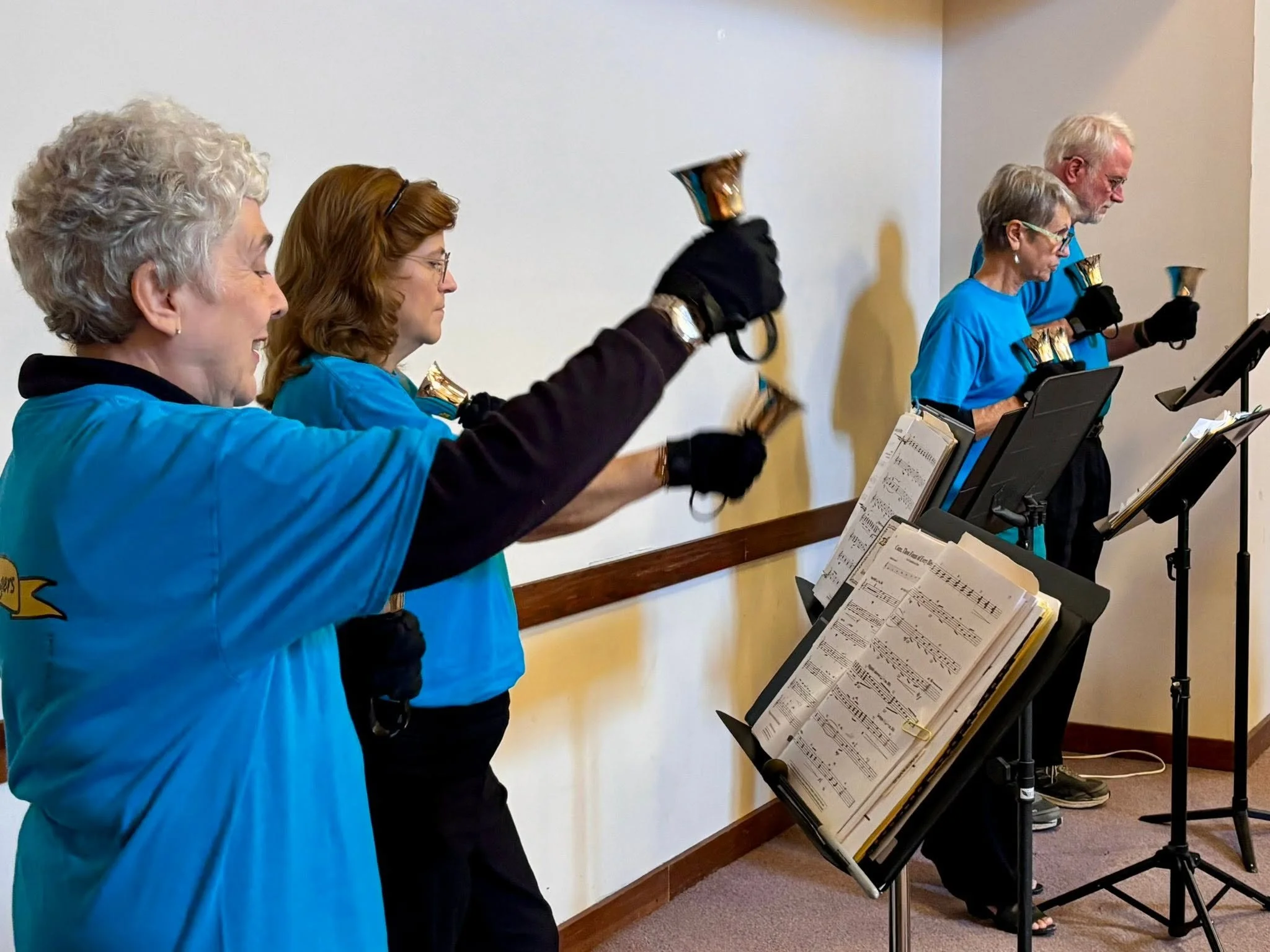 A group of five elderly people standing and playing bells, reading sheet music from music stands in front of them, dressed in matching blue shirts, set against an indoor plain white wall.