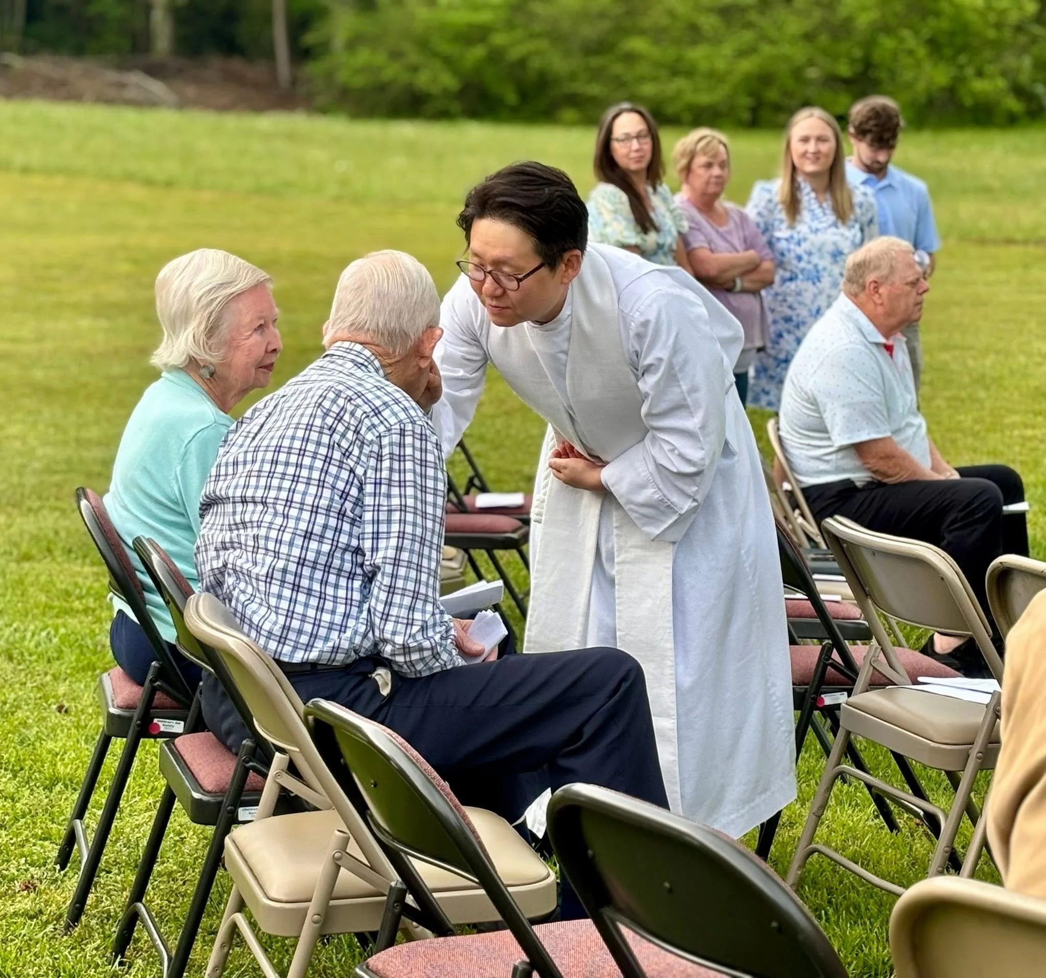 Group of elderly people sitting outdoors in chairs, while a man dressed as a priest or spiritual leader talks to a woman in a mint green top. Several women and a man stand in the background observing. The setting appears to be a garden or park with grass and trees.