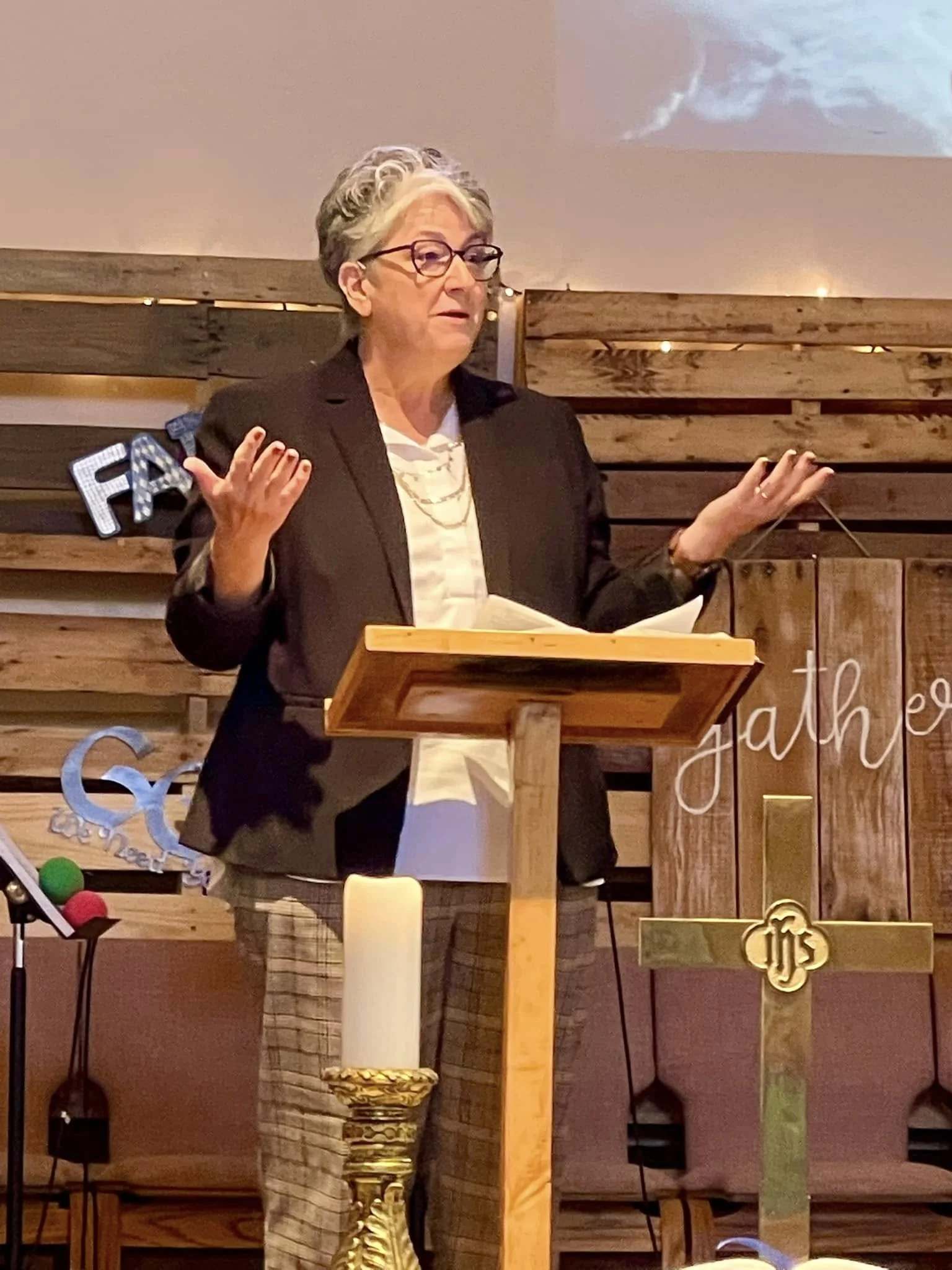 A woman with short gray hair and glasses standing at a wooden lectern, speaking and gesturing with her hands, inside a room with wood-paneled walls and a decorative candle and gold ornament.
