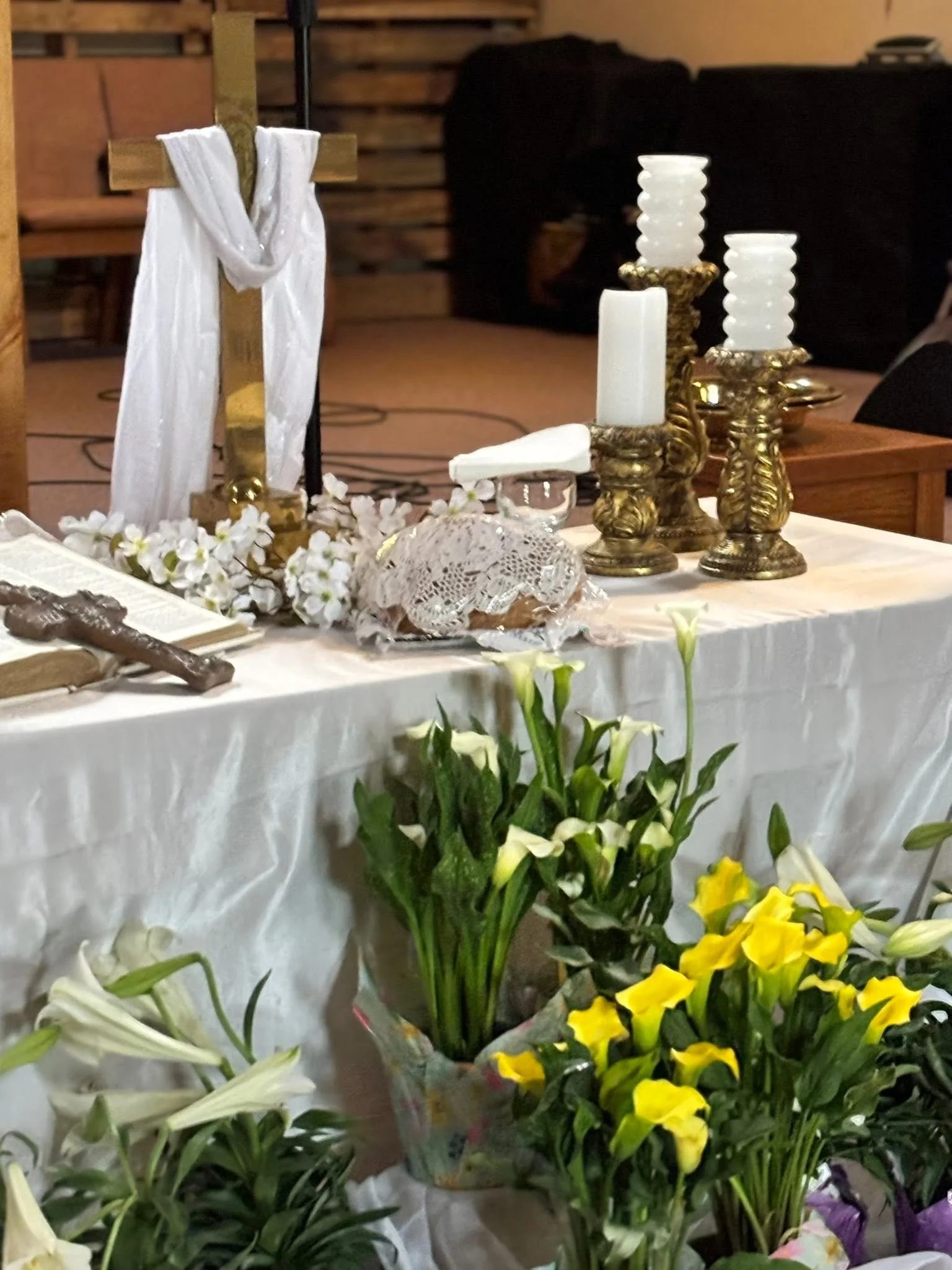 Religious altar with a wooden cross draped with white cloth, white and gold candles, and floral arrangements including white, yellow, and purple flowers.