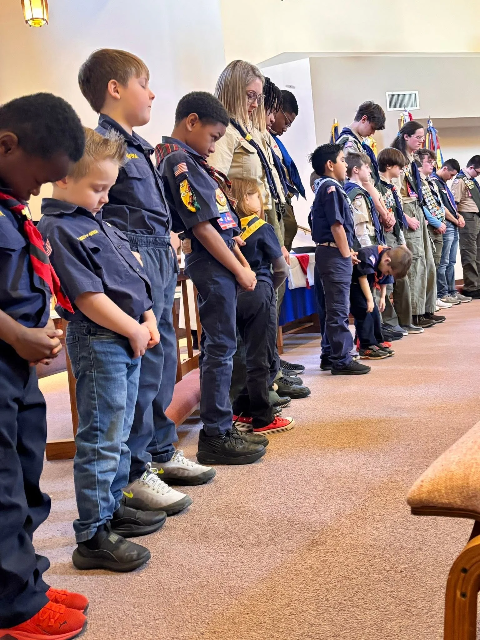 A line of young boy and girl scouts standing with heads bowed and eyes closed inside a room with beige walls and carpeted floor. Some possibly have their hands clasped in front of them or at their sides, participating in a moment of prayer or reflection.