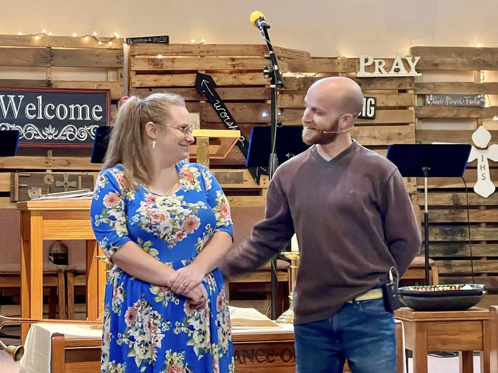 A woman and a man are holding hands and smiling at each other in a church or community setting with rustic wooden decor and framed signs that read 'Welcome' and 'Pray' in the background.