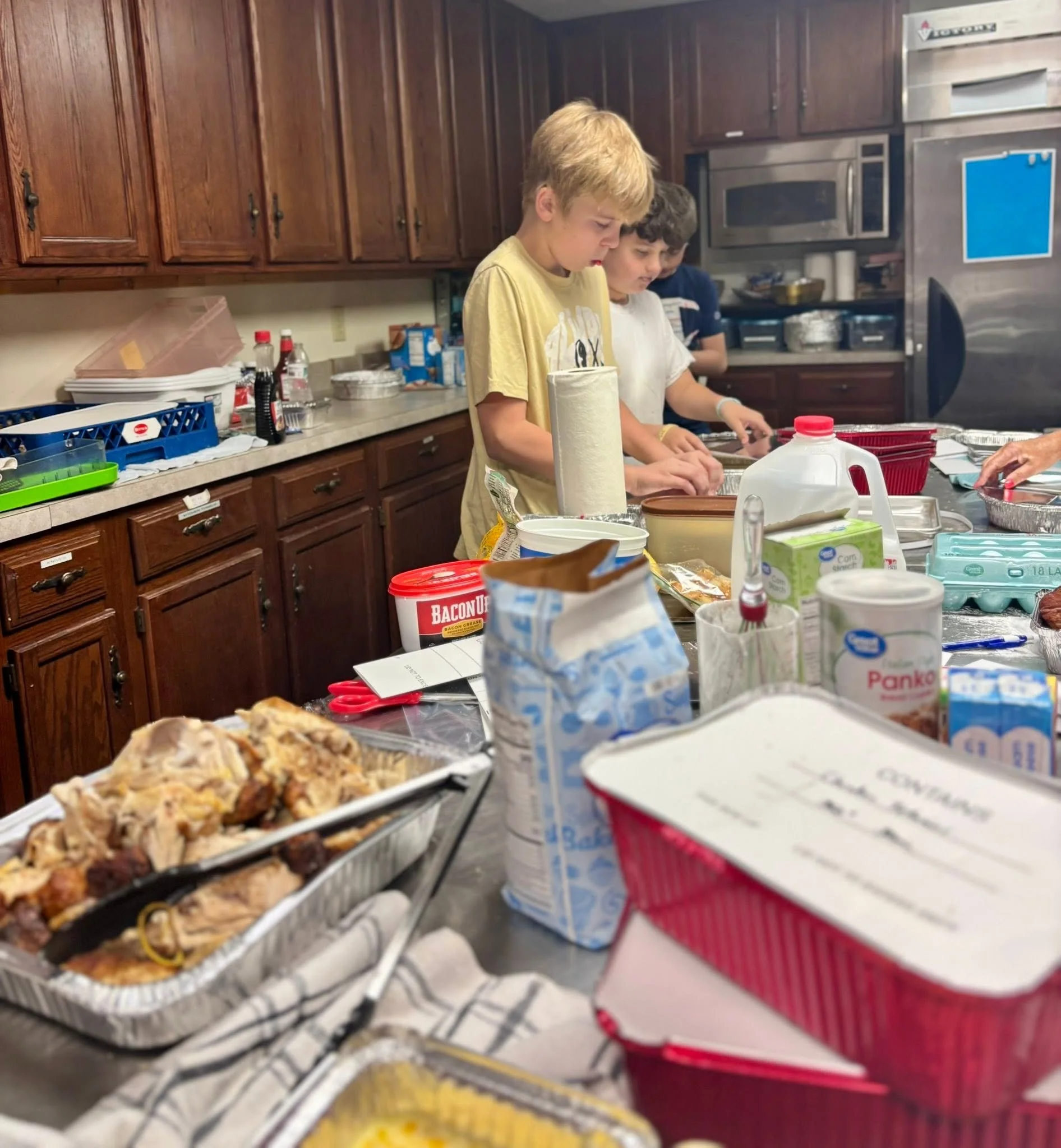Children making pizza in a kitchen with wooden cabinets and various ingredients on the counter.