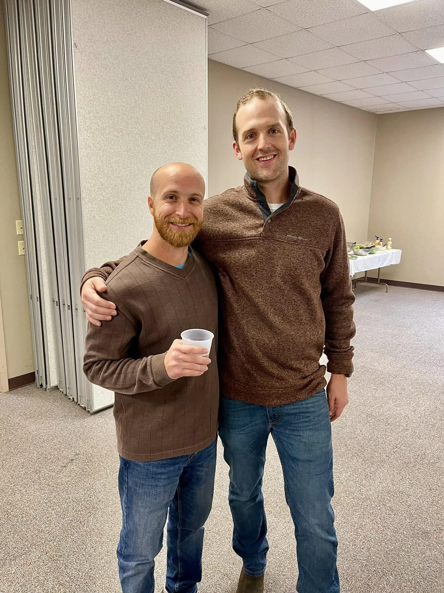 Two smiling men standing in an indoor room, with one man holding a plastic cup. The man on the left has a beard, bald head, and is wearing a brownish sweater. The man on the right has short hair, no beard, and is wearing a brown quarter-zip sweater and jeans.