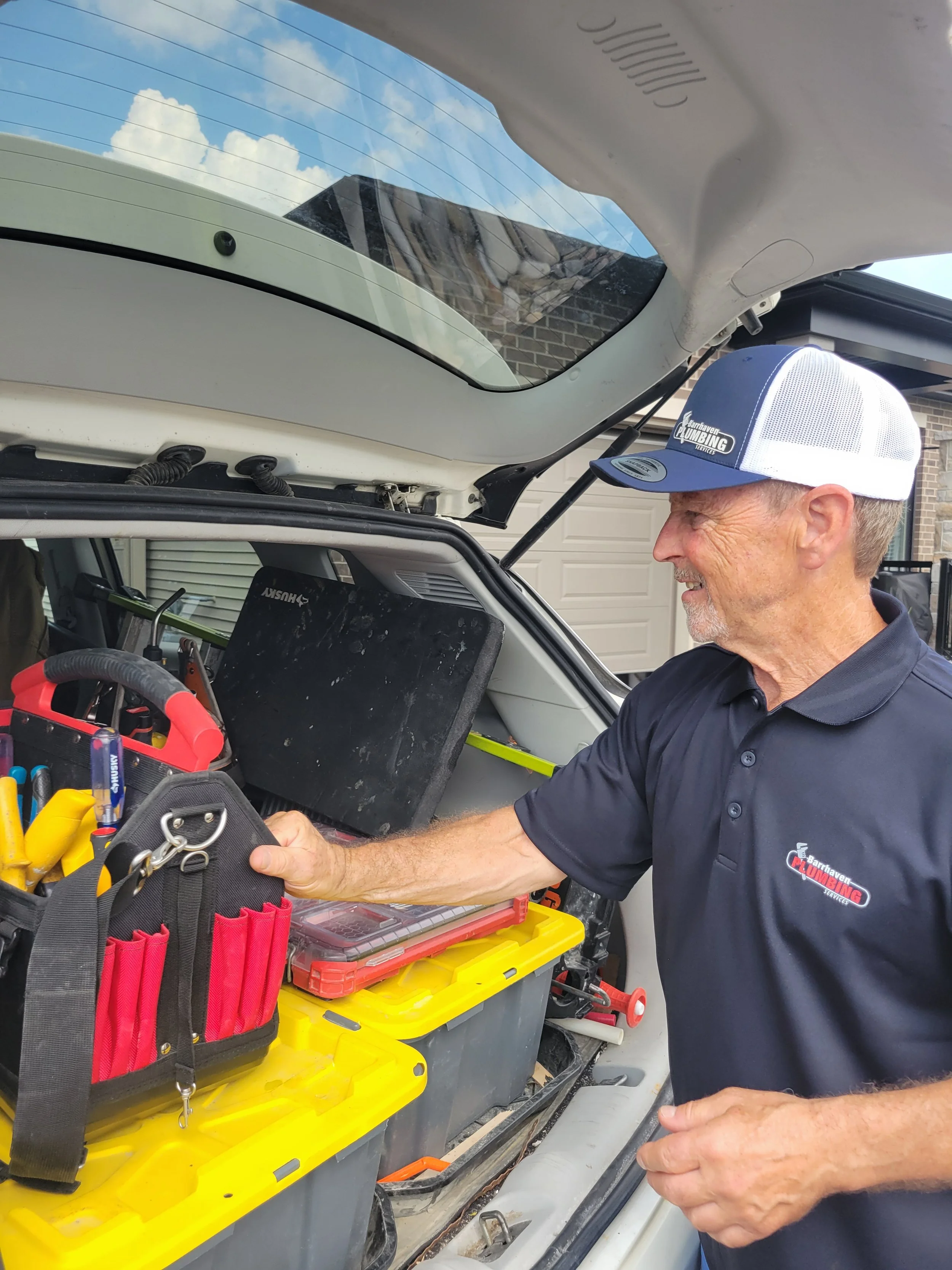 A man in a navy polo shirt and a trucker cap is working on a vehicle, with an open trunk filled with tools and equipment, including a red tool pouch and yellow containers. The background shows a garage with a closed white door.