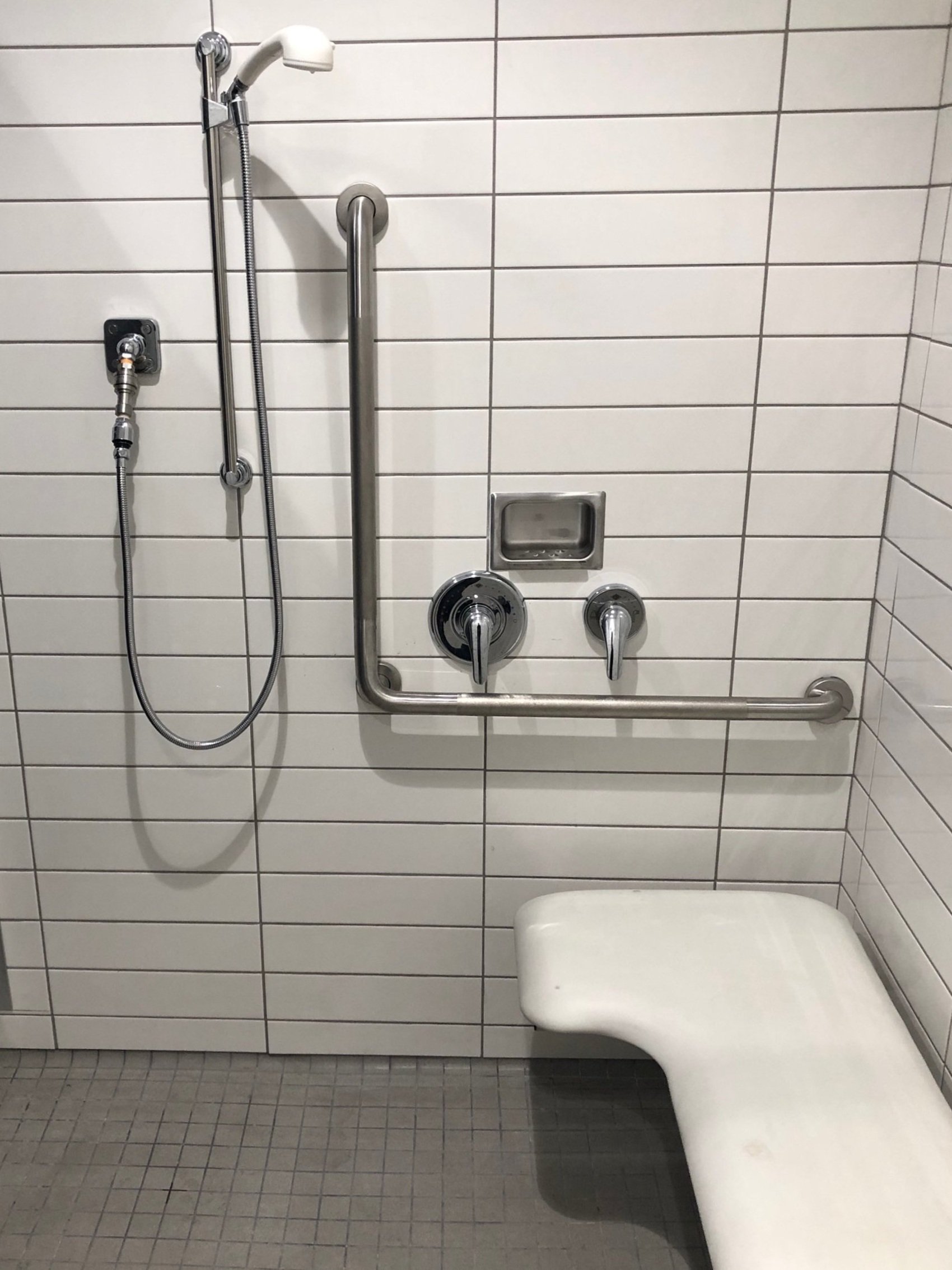 Accessible shower area with a wall-mounted head, handrails, a soap dish, and a white shower chair, with tiled walls and floor.