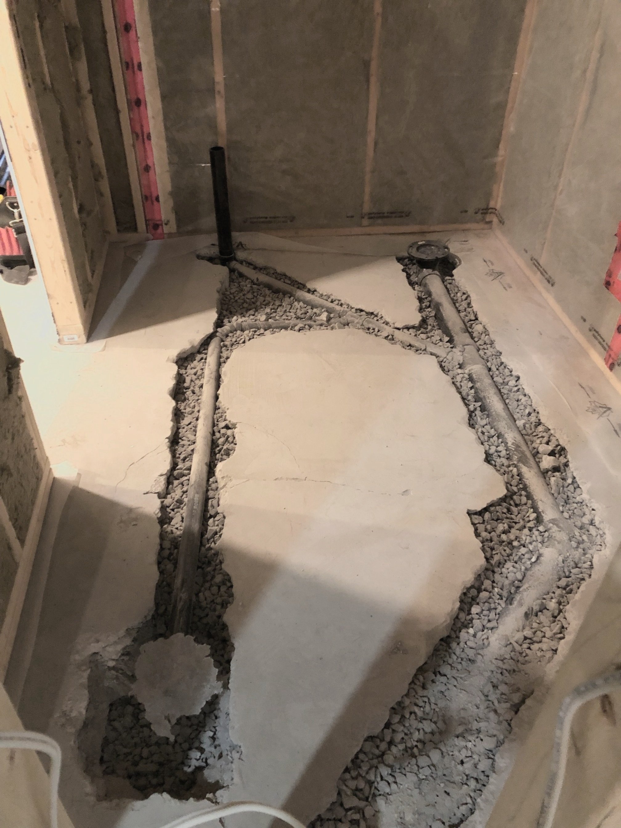 A bathroom under construction with exposed pipes and framing, a large piece of broken concrete, gravel surrounding the pipes, and unfinished wall surfaces.
