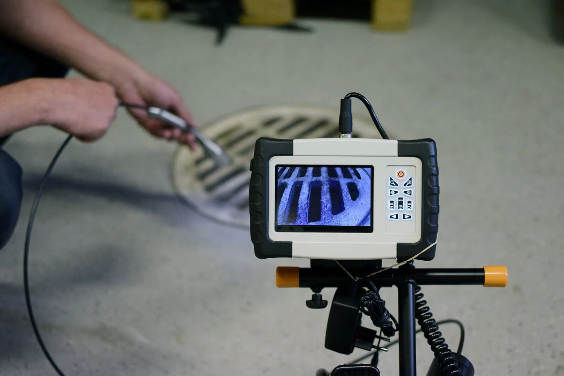 A pipe inspection camera with a monitor displaying an image of a drain pipe, positioned near a floor drain with a person holding a tool or cable nearby