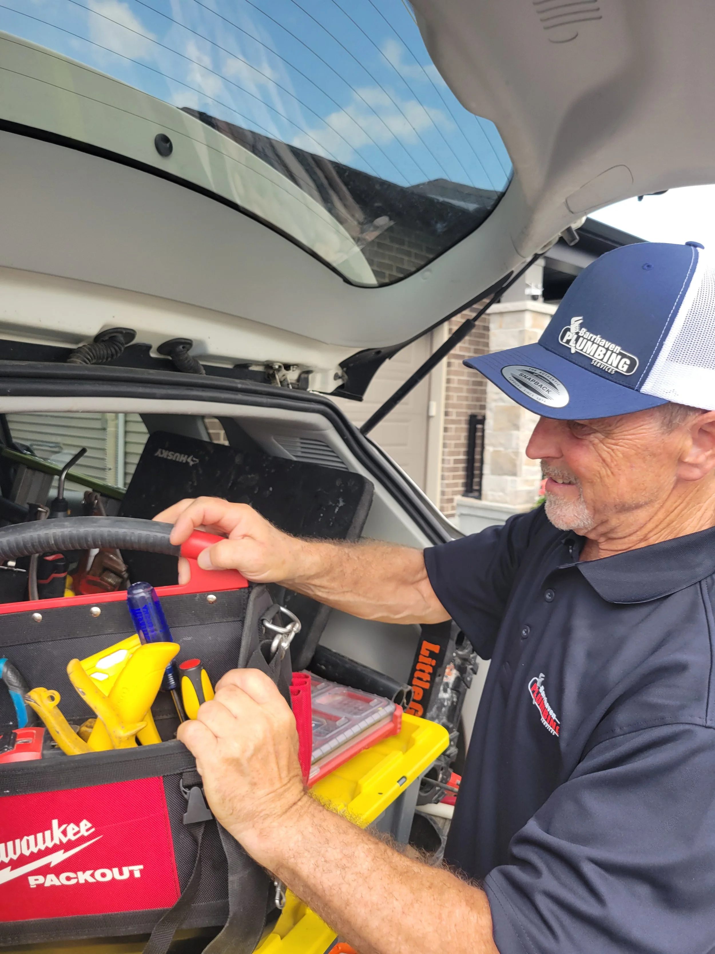 A man wearing a blue cap and a black shirt is working on tools in an open toolbox attached to a vehicle. He is outdoors near brick buildings and a garage with a car and various tools inside.