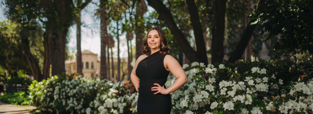 A woman in a black dress standing outdoors beside white flowering bushes, with trees and a building in the background.