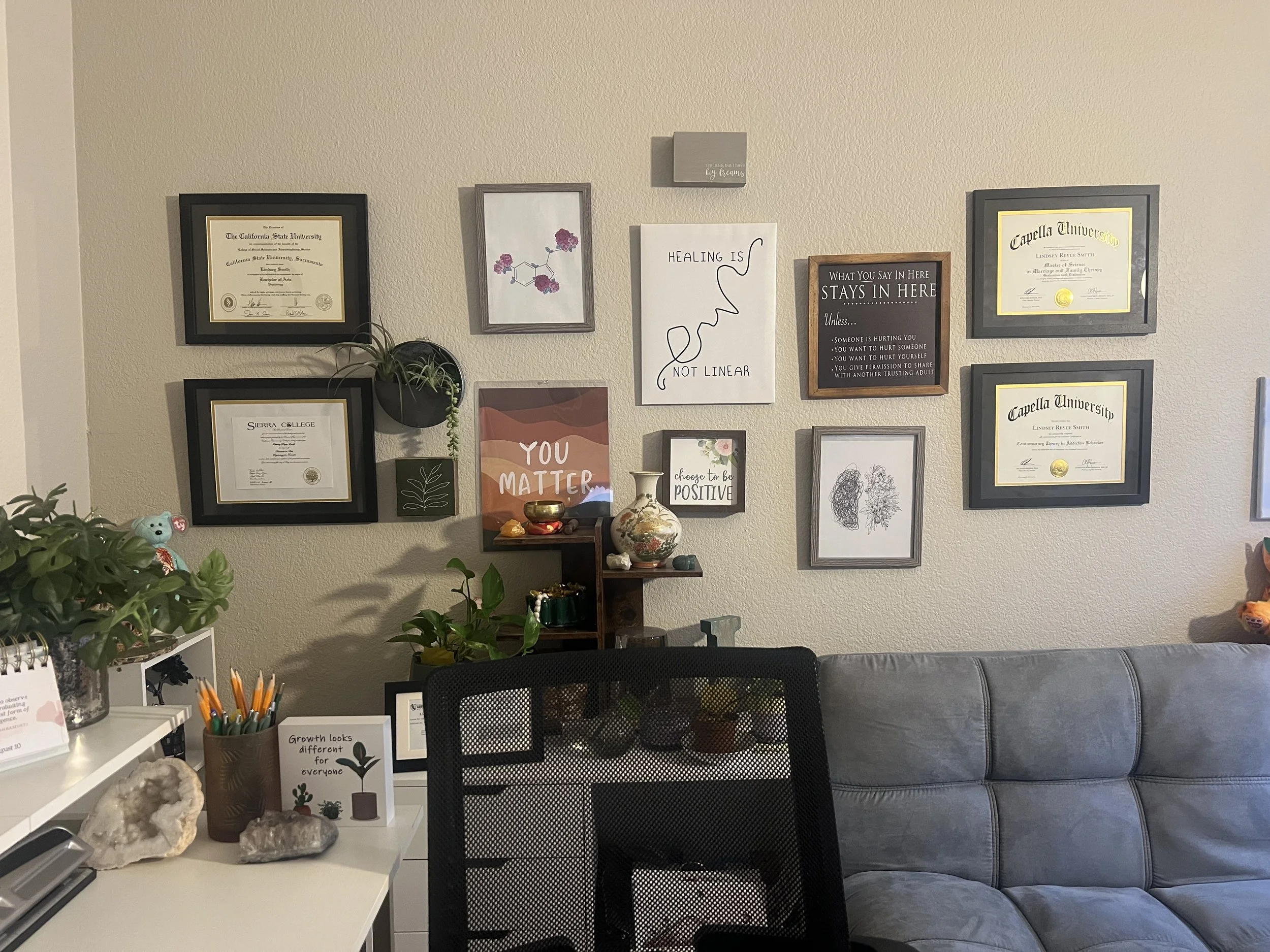 Office space with wall display of framed certificates, motivational signs, and artwork. A small wooden shelf holds plants and decorative items. In the foreground, a white desk has office supplies, a plant, and rocks. A phone and a sign about growth are on the desk. A gray sofa is on the right side.
