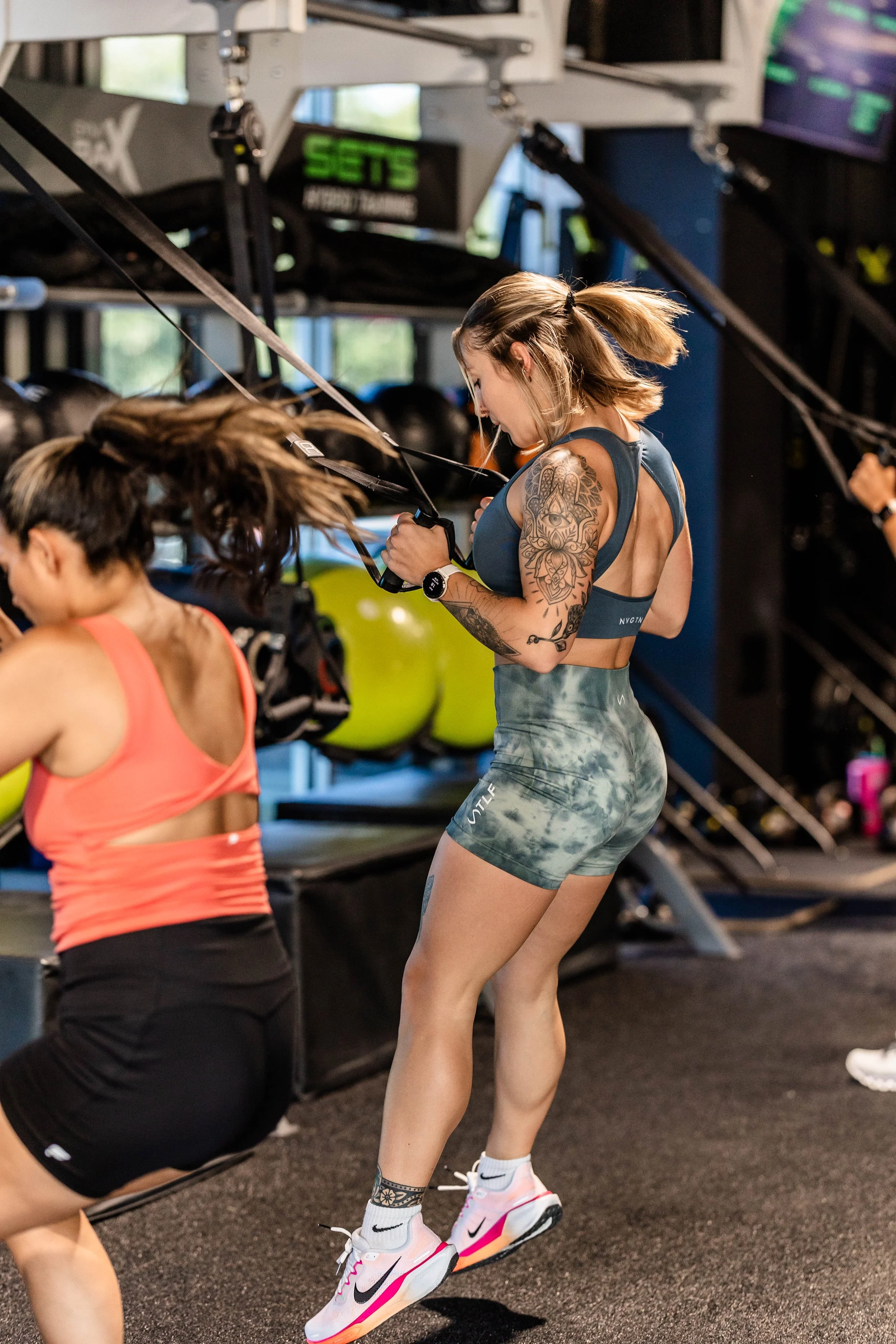 A woman with tattoos on her arm and leg, wearing athletic clothing, earbuds, and Nike sneakers, working out at a gym with rowing machines and weights.