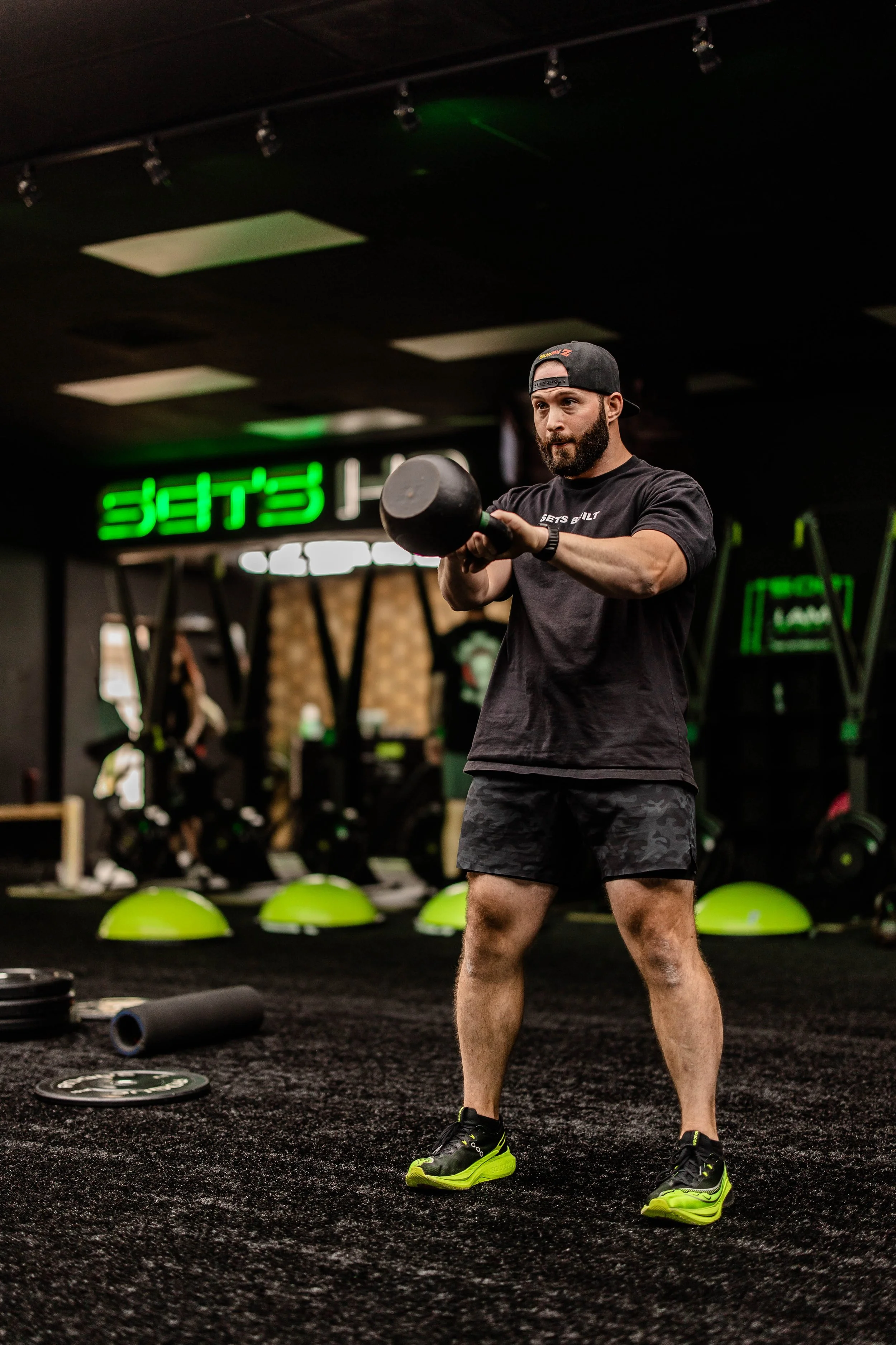 A man with a beard wearing a black cap and black t-shirt lifting a kettlebell during a workout in a gym. The gym has black flooring, green fitness equipment, and neon signs in the background.