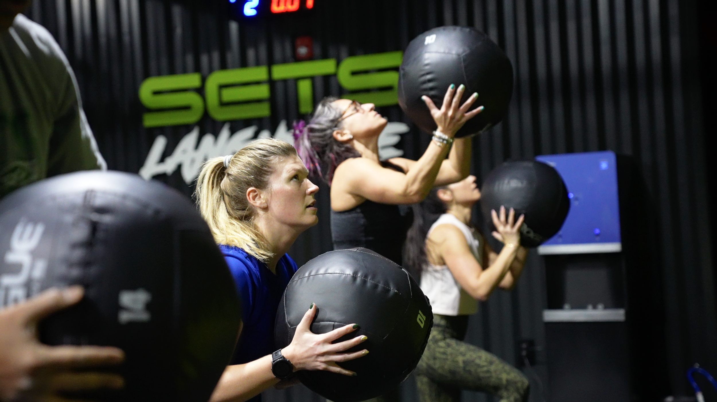 People participating in a fitness class using black medicine balls with a neon green sign reading 'SENS' in the background.