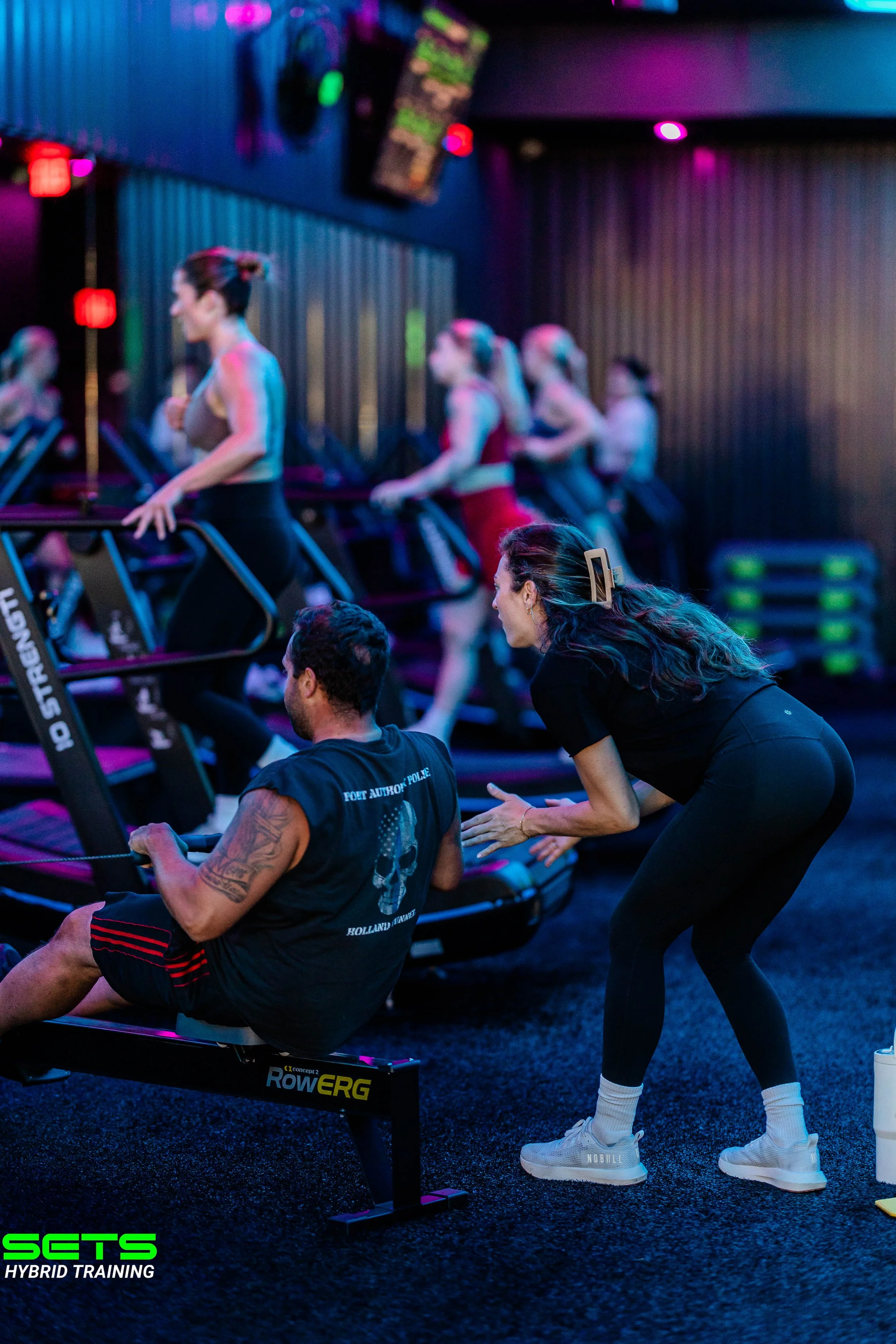 Women in athletic attire attending a fitness class on rowing machines in a dimly lit gym with colorful neon lighting and a trainer guiding them.