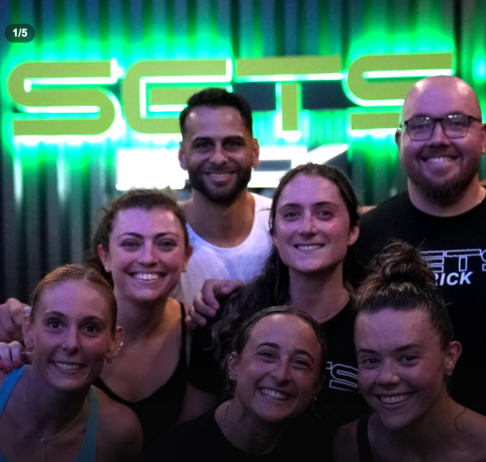 Group of seven smiling people posing in front of a green neon sign that reads 'SCTE' at night or in a dark setting.