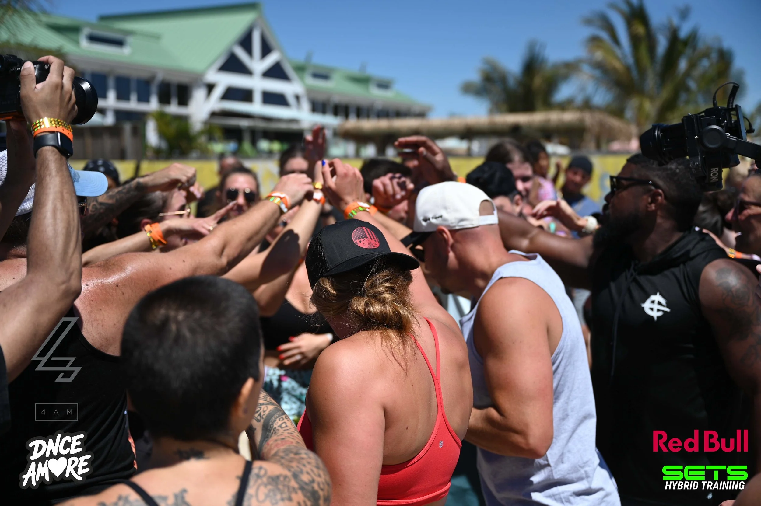 A large crowd of people gathered outdoors on a sunny day, participating in a group event or celebration, with some wearing hats and sunglasses. There is a building with a green roof in the background, along with trees. The image has logos promoting Red Bull, dance, and fitness.