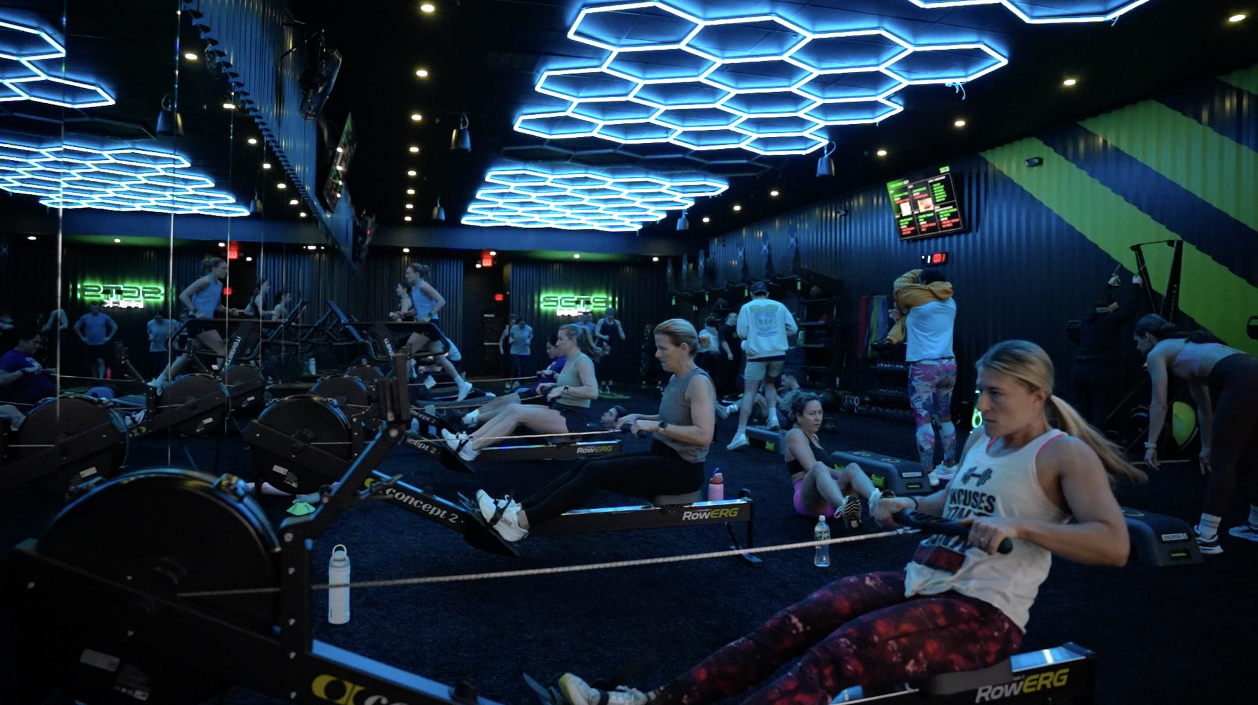 People participating in an indoor rowing class with neon blue lighting and digital screens on the walls.